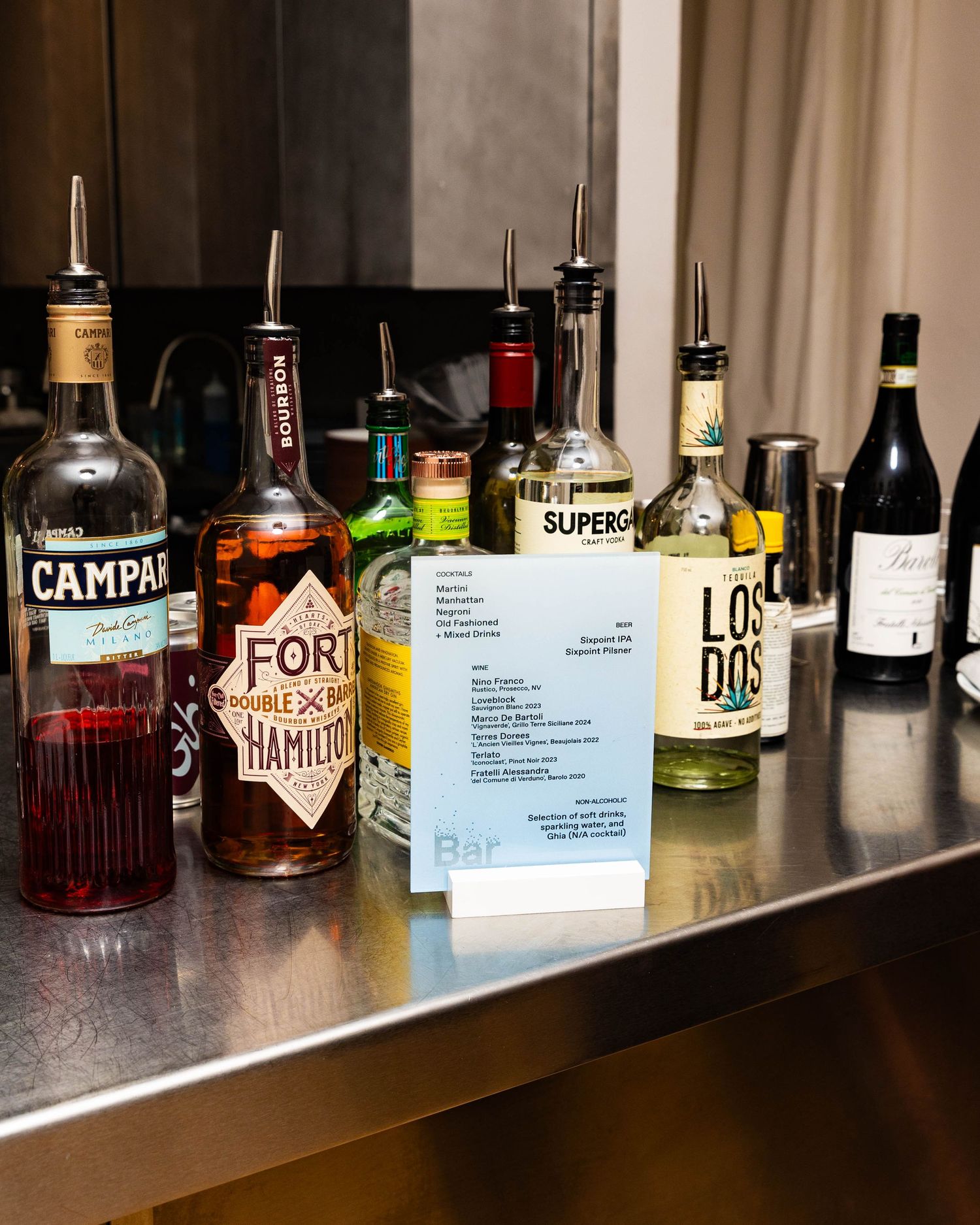 Bottles of Campari, Fort Hamilton, Superga, Los Dos and red wine line a stainless steel bar next to a small drink menu card.