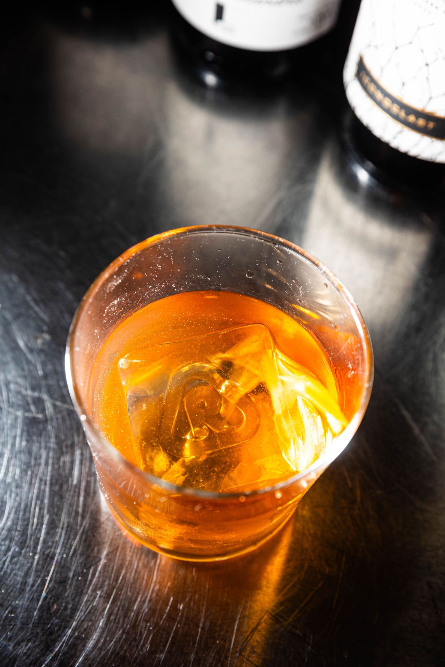 Amber cocktail with a large ice cube in a rocks glass on a brushed metal bar top, with liquor bottles blurred in the background.
