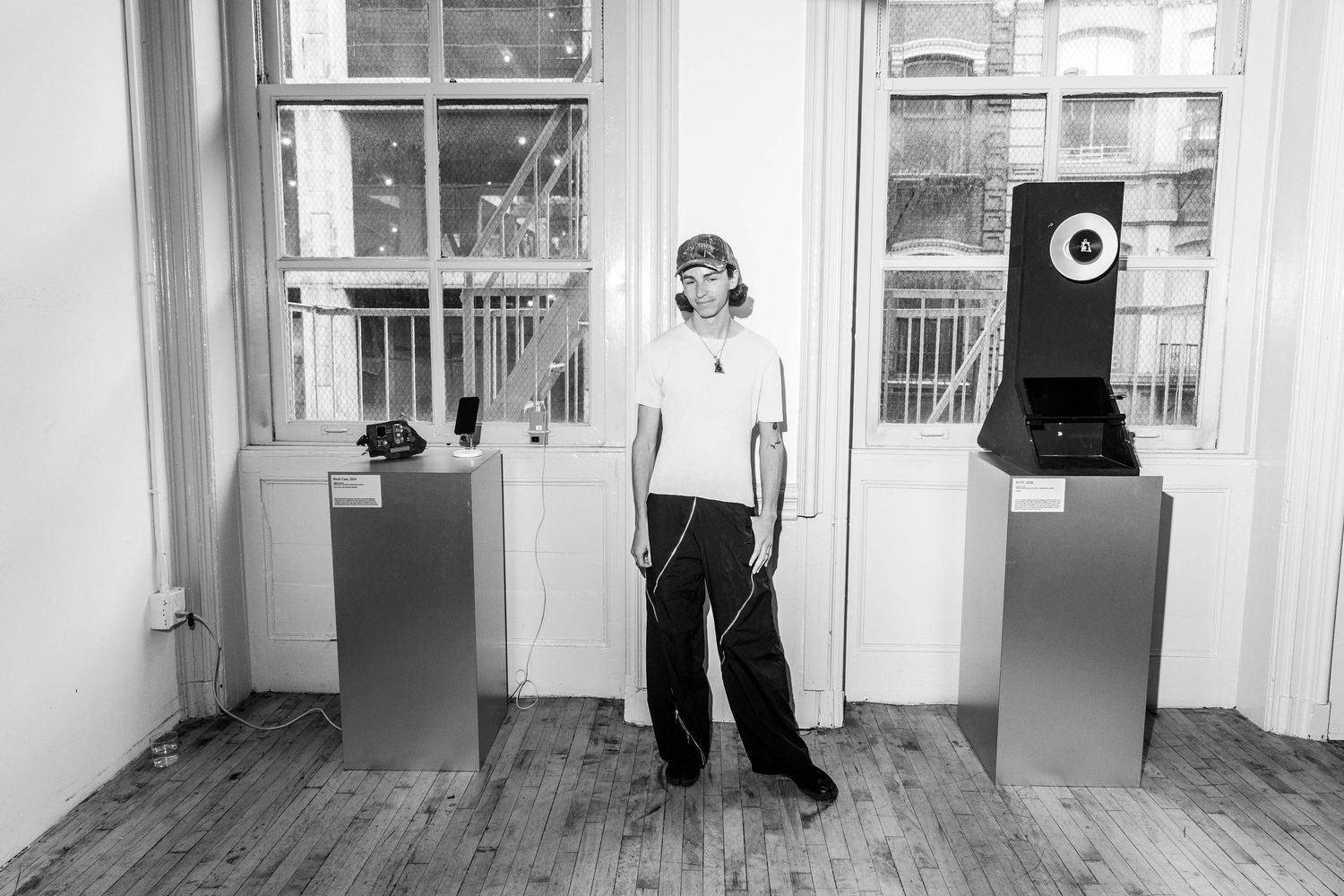 A person in a white t-shirt and dark pants stands between two metal pedestals displaying vintage phone equipment in a sunlit loft gallery.