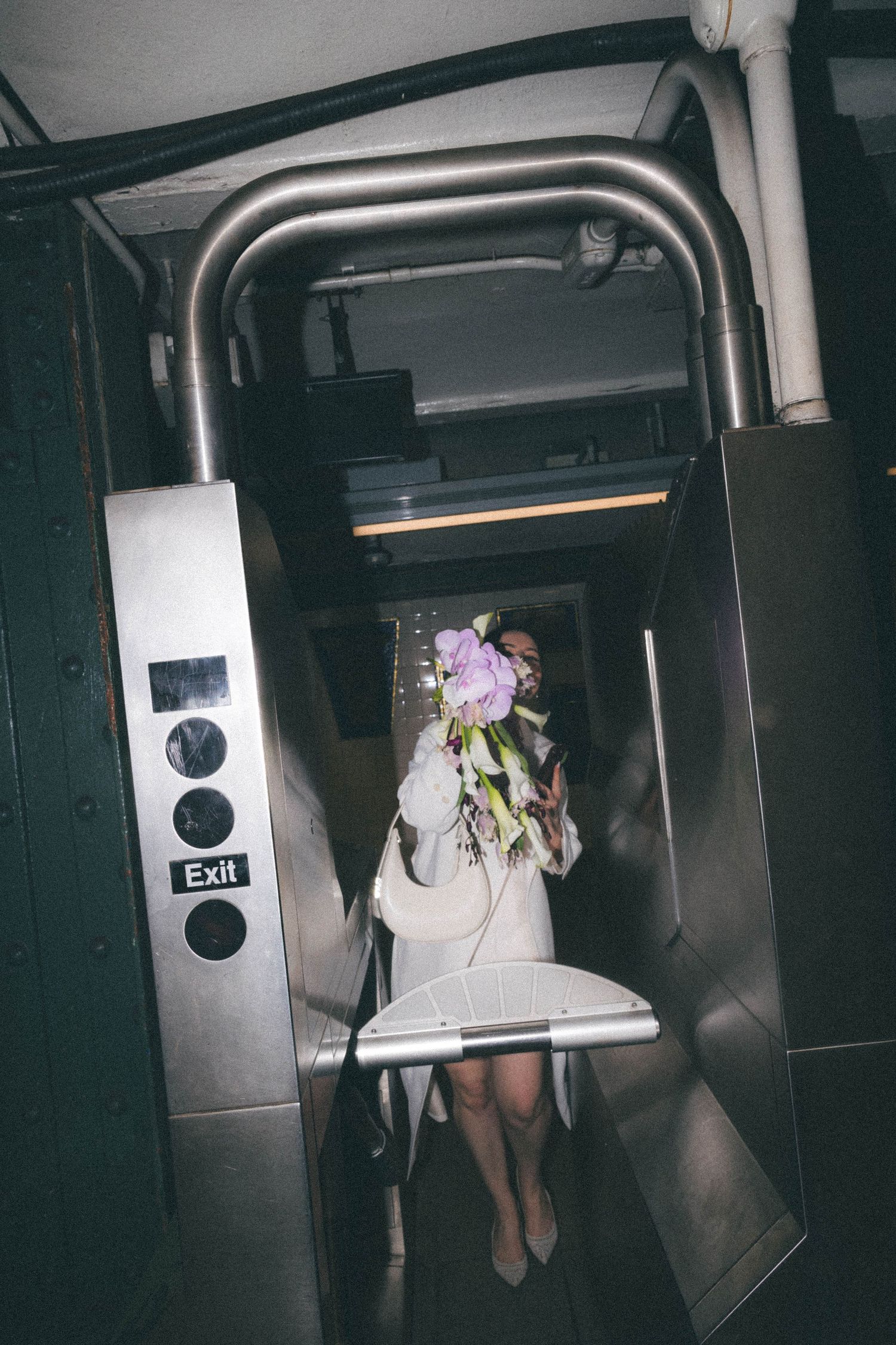 Bride in white dress holds bouquet over face while exiting NYC subway turnstile, flashing peace sign.