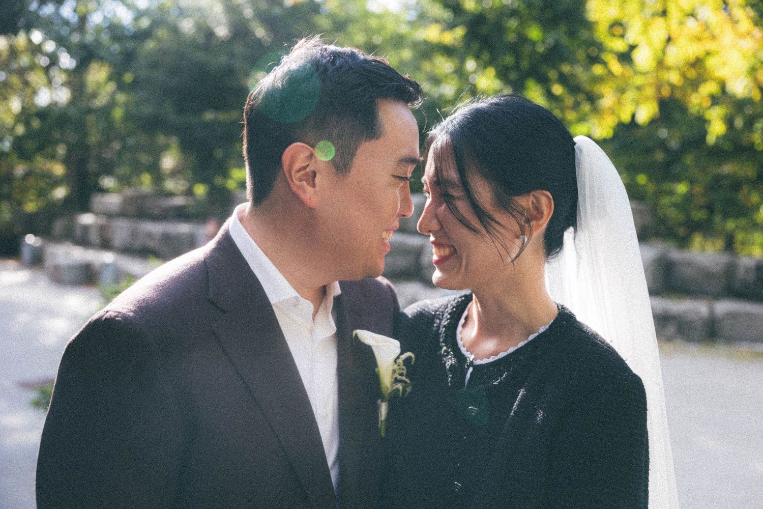 Bride and groom share intimate moment touching foreheads in sunlit outdoor wedding portrait.