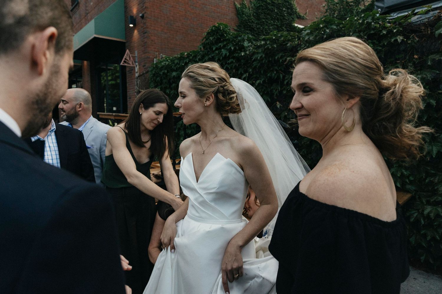 Bride in elegant white gown with veil greets guests outdoors at wedding reception with bridesmaids nearby.