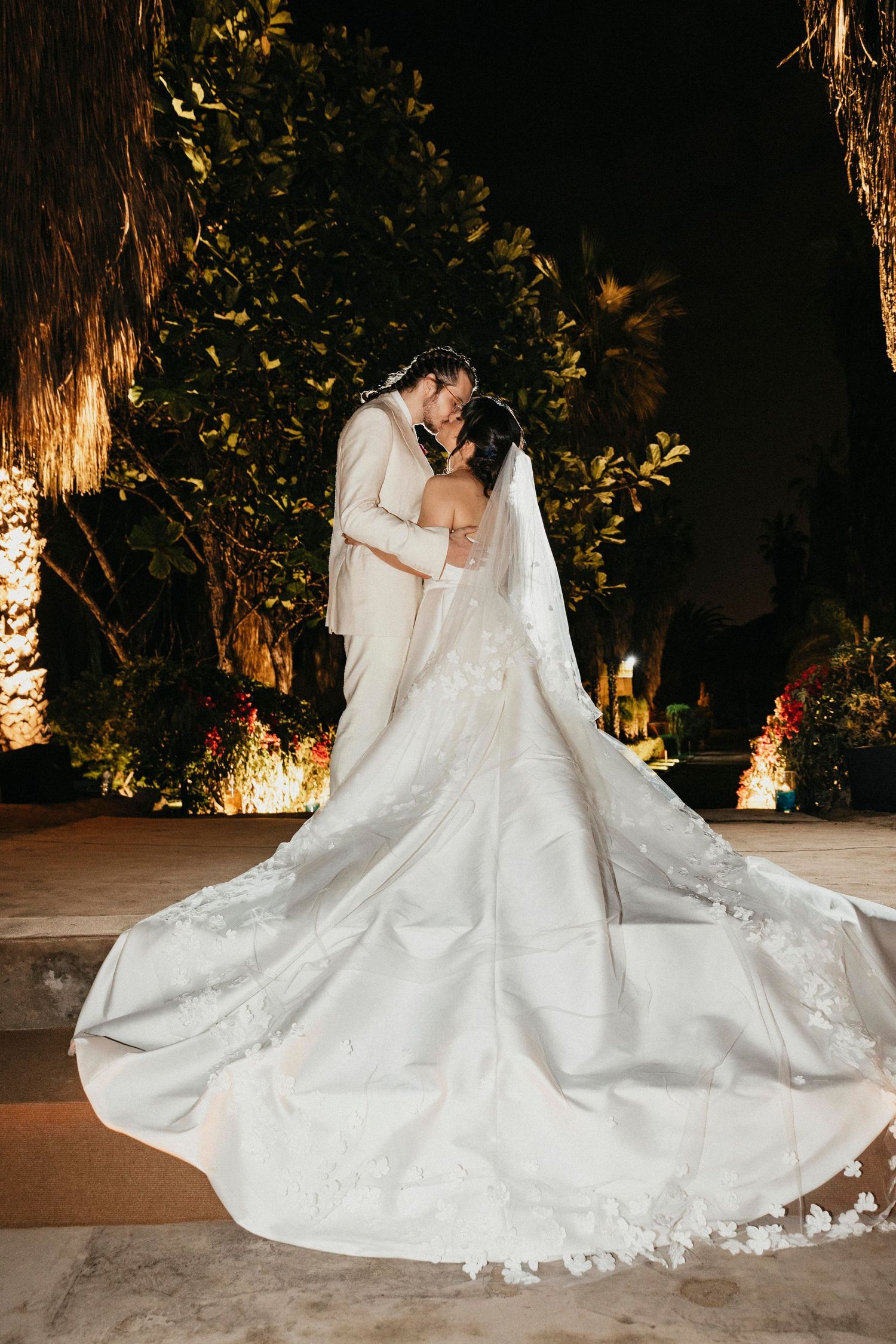 Bride and groom share a kiss as her lace cathedral veil fans dramatically across the ground at night.