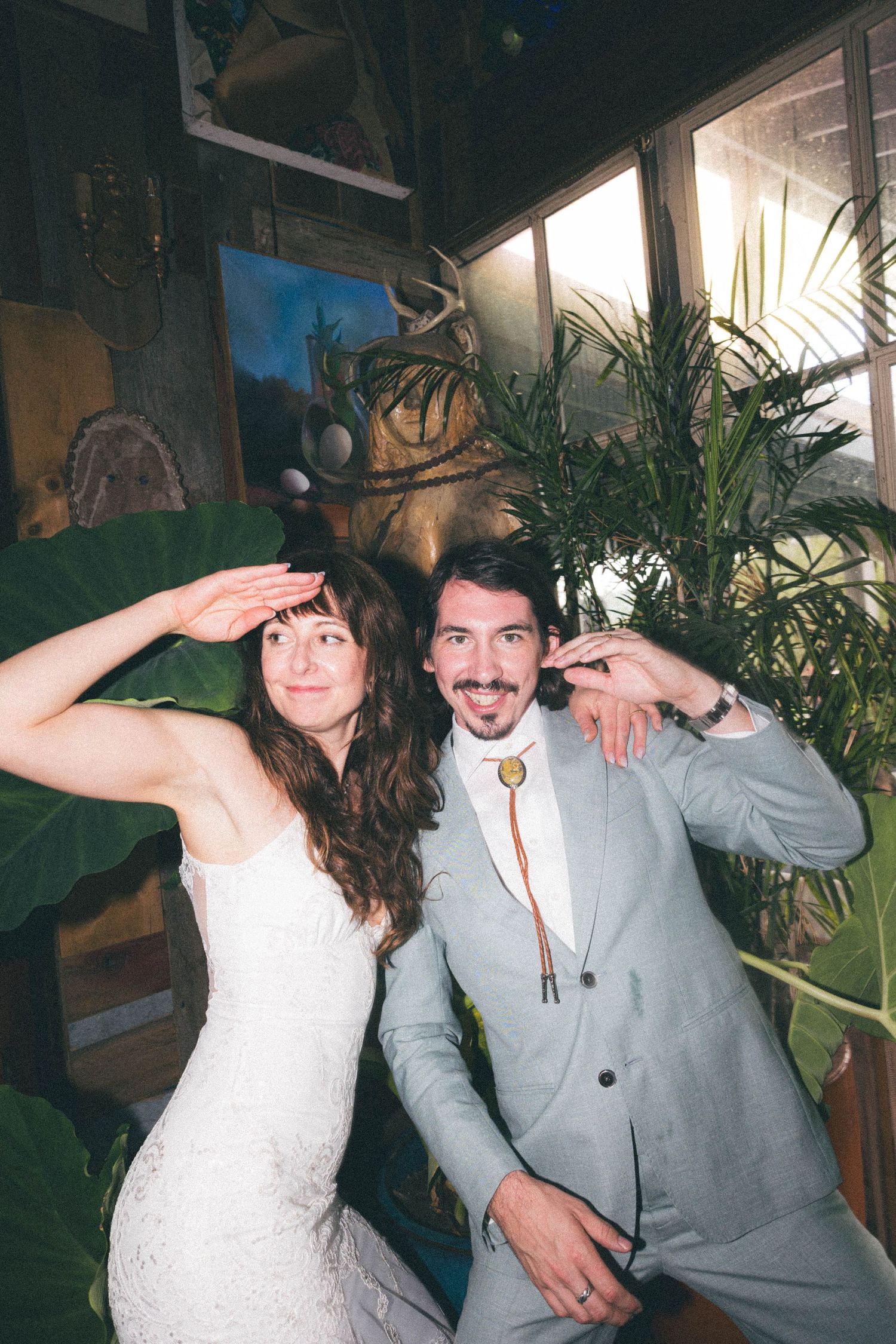 Playful bride and groom pose together saluting in a rustic venue decorated with tropical plants.
