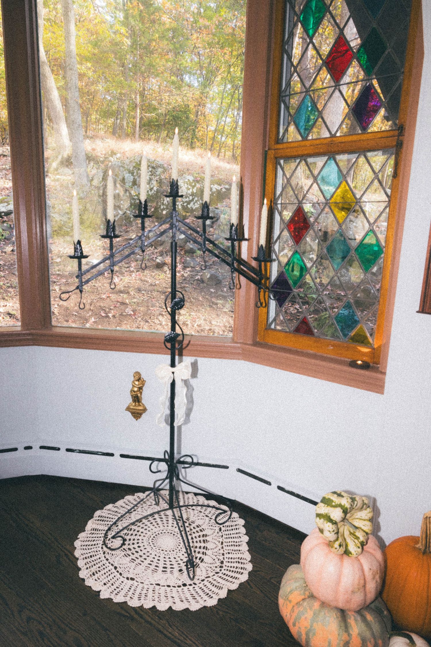 Elegant black iron candelabra on lace doily near colorful stained glass bay window with autumn pumpkins.
