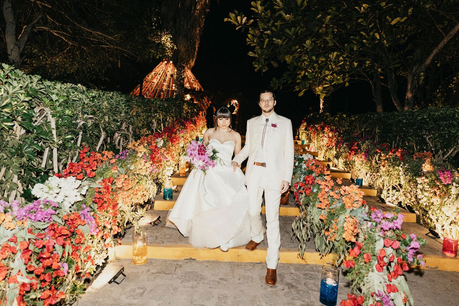 Bride and groom walk hand in hand through a vibrant floral-lined pathway at their outdoor evening wedding.