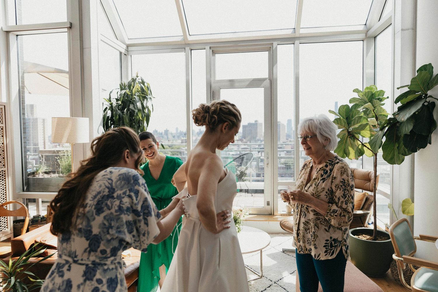 Bride in white gown is assisted by family and friends in a bright sunlit penthouse getting-ready suite.