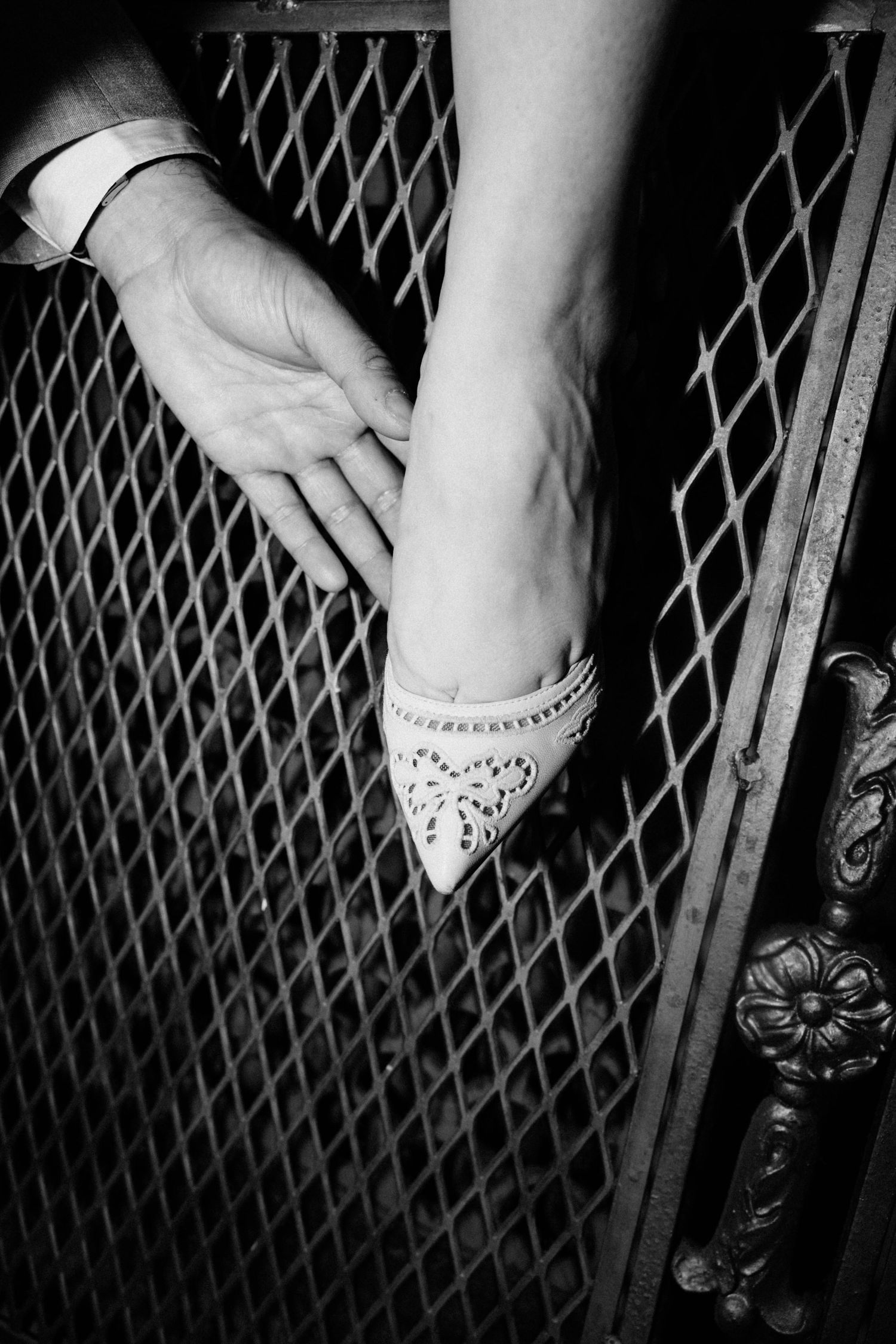 Close-up black and white photo of bride and groom holding hands on ornate metal gate.
