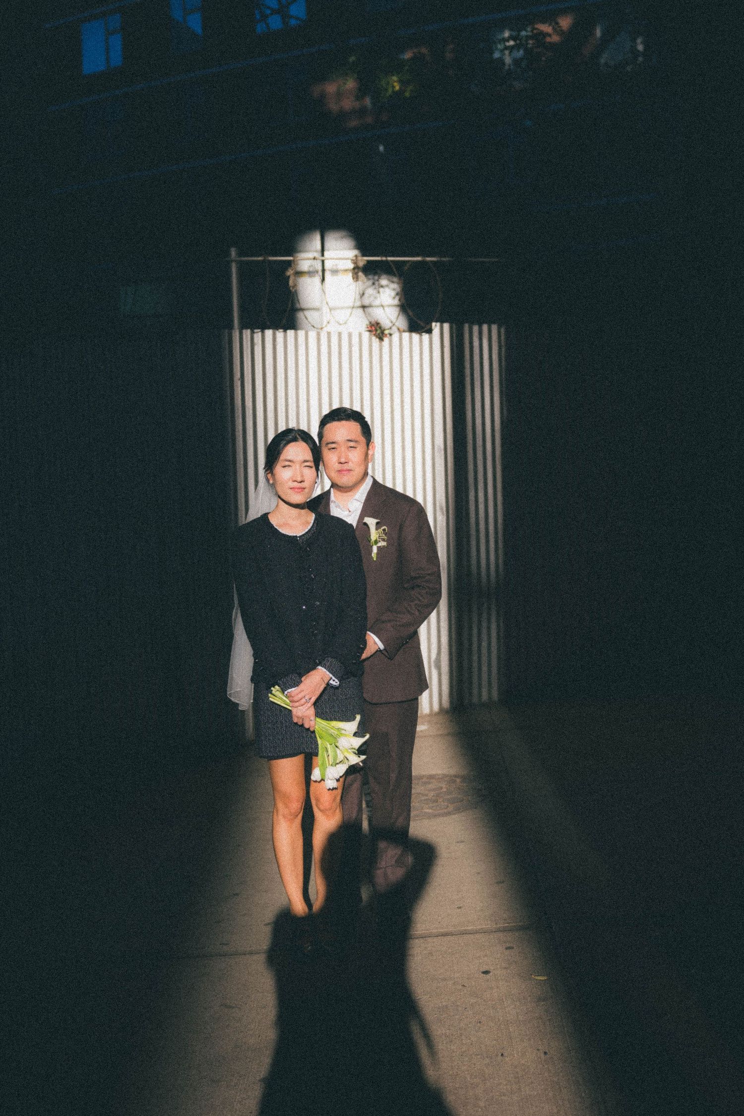 Couple poses in dramatic spotlight at night, woman holding white calla lilies, man in brown suit with boutonniere.