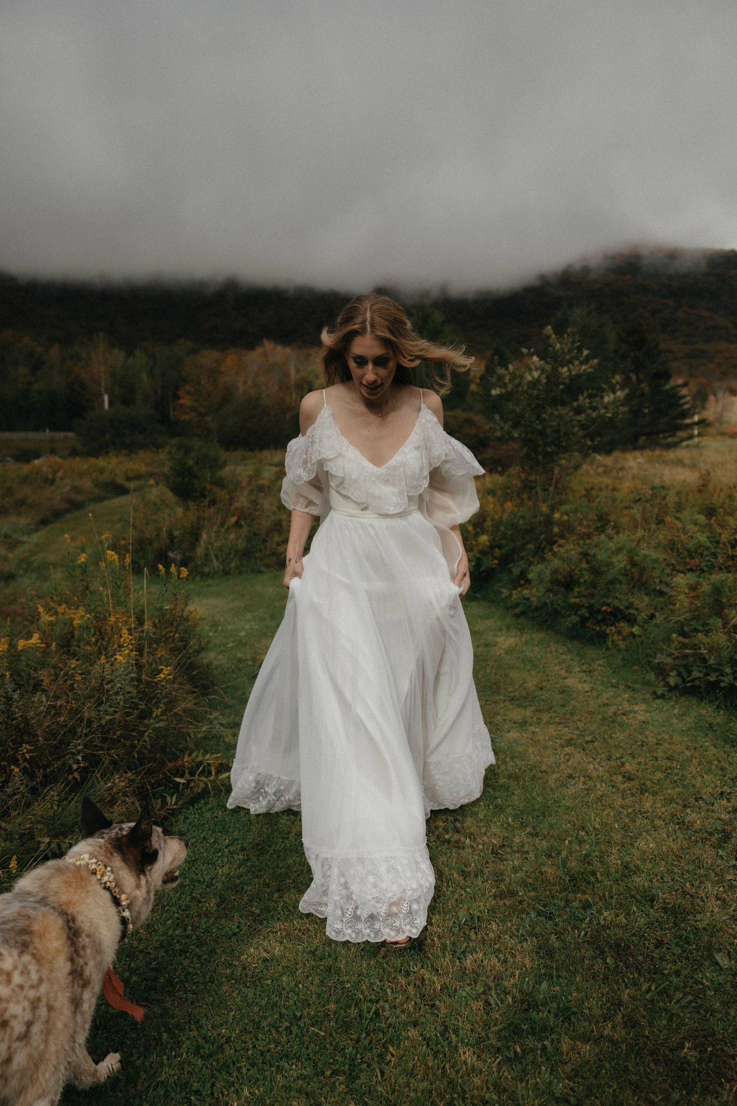 Bride in flowing white lace gown walking with dog across green meadow on overcast day.