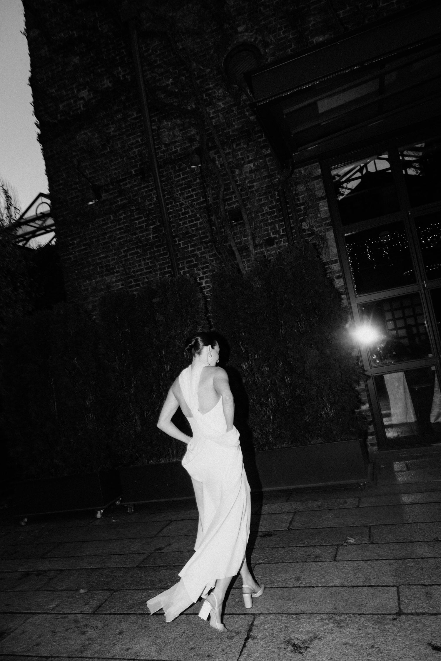 Bride in elegant backless white gown posing outside ivy-covered brick building at night.