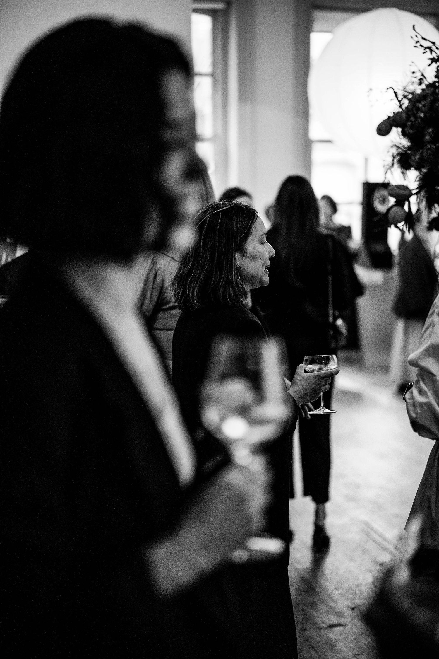 Black and white photo of guests holding wine glasses and chatting at an indoor gathering, with a blurred woman in the foreground.