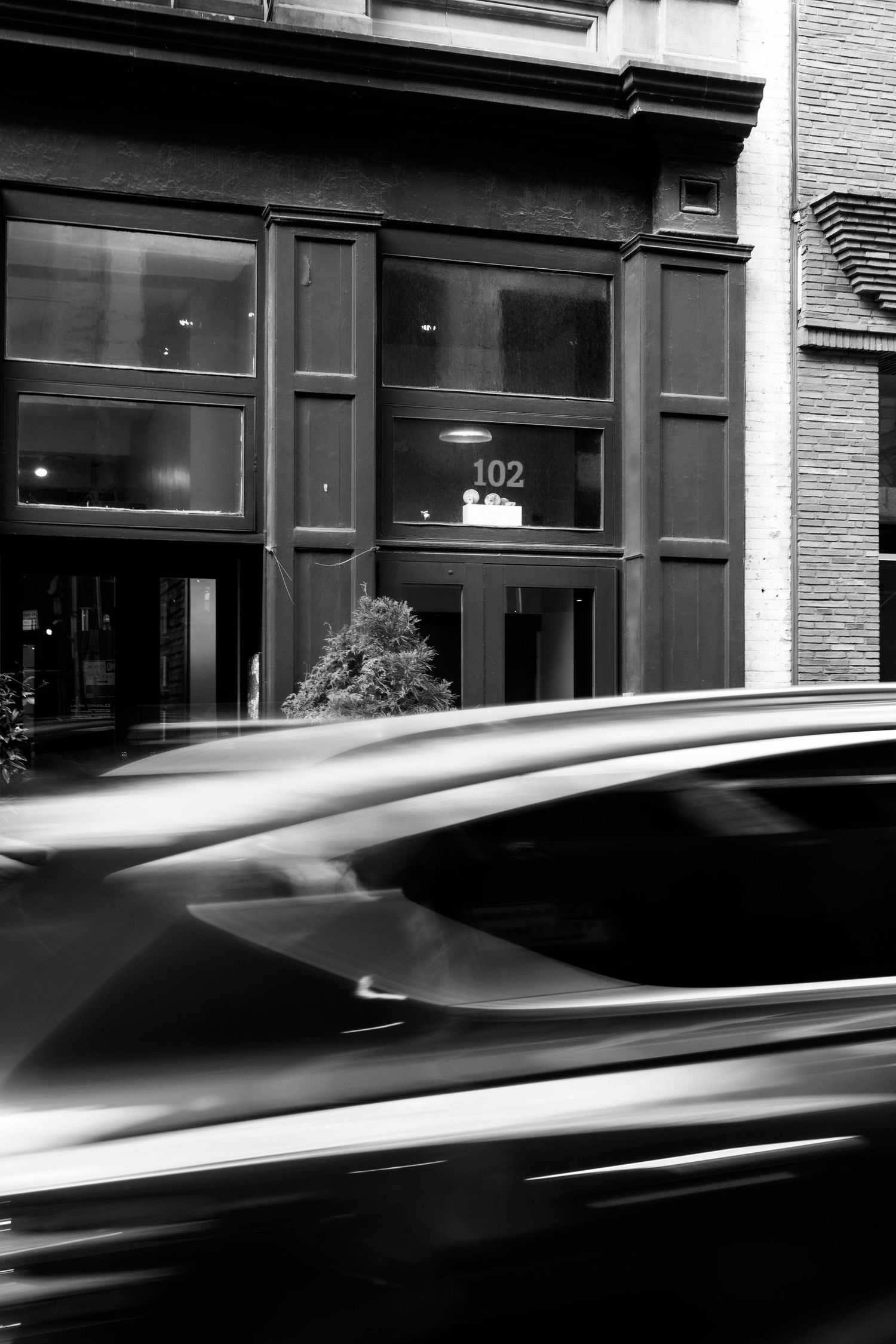 Black and white photo of a blurred car speeding past a historic building entrance marked 102 with a small shrub out front.