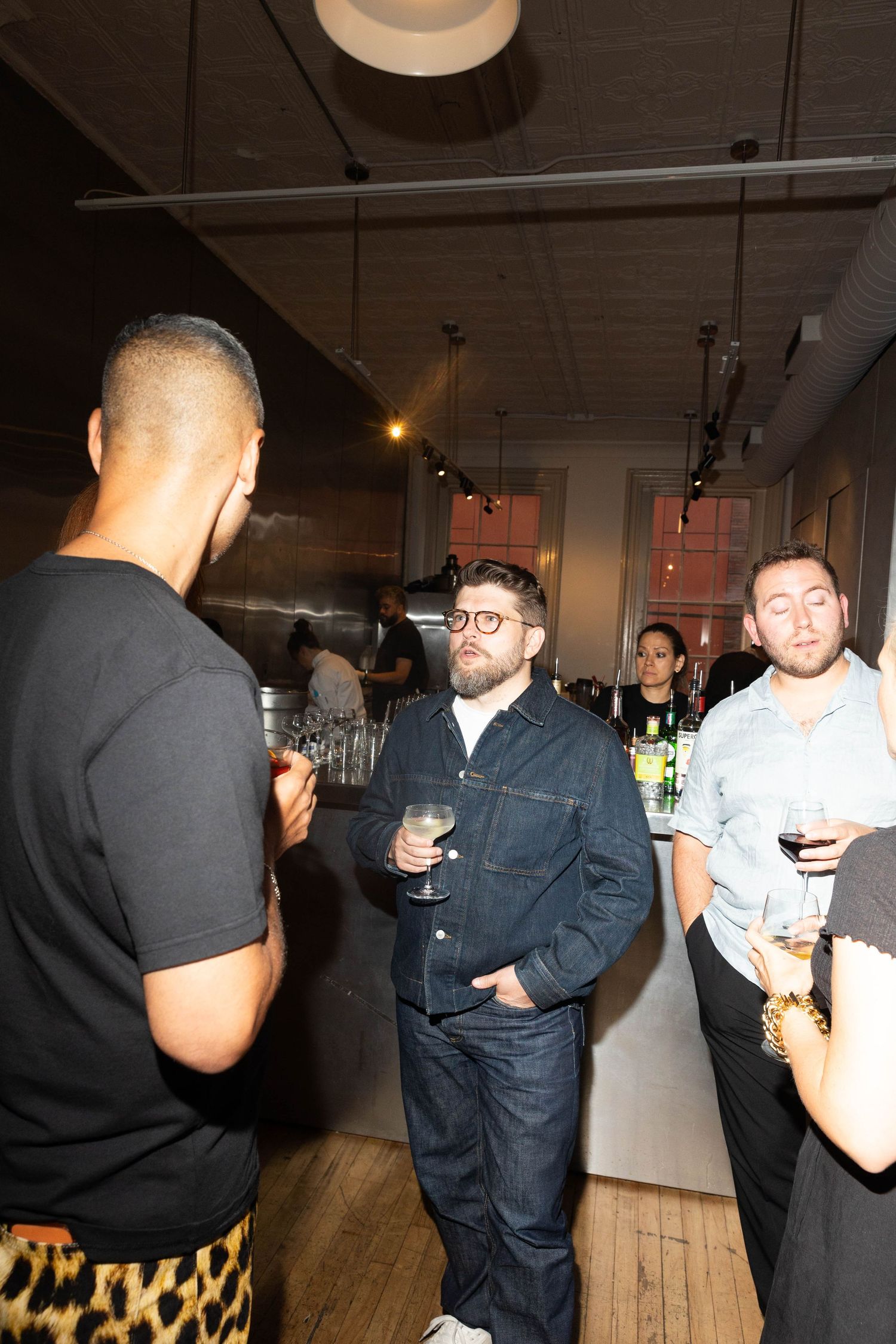 Guests in casual outfits hold wine glasses and chat at a stylish indoor event with a bar and bartenders in the background.