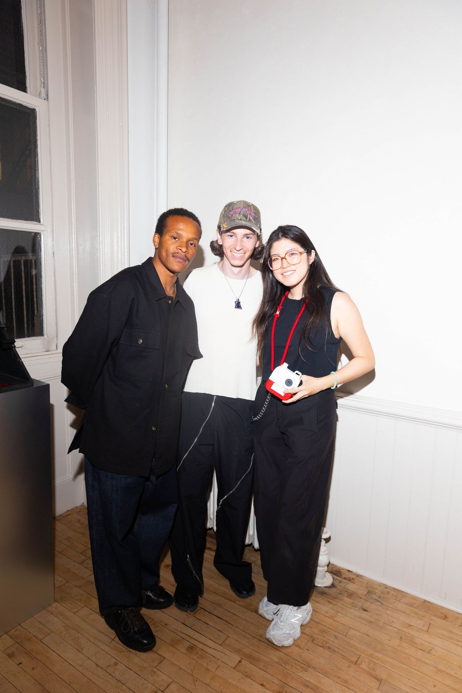 Three smiling people pose together in a bright room with wood flooring, one holding a small red and white camera.