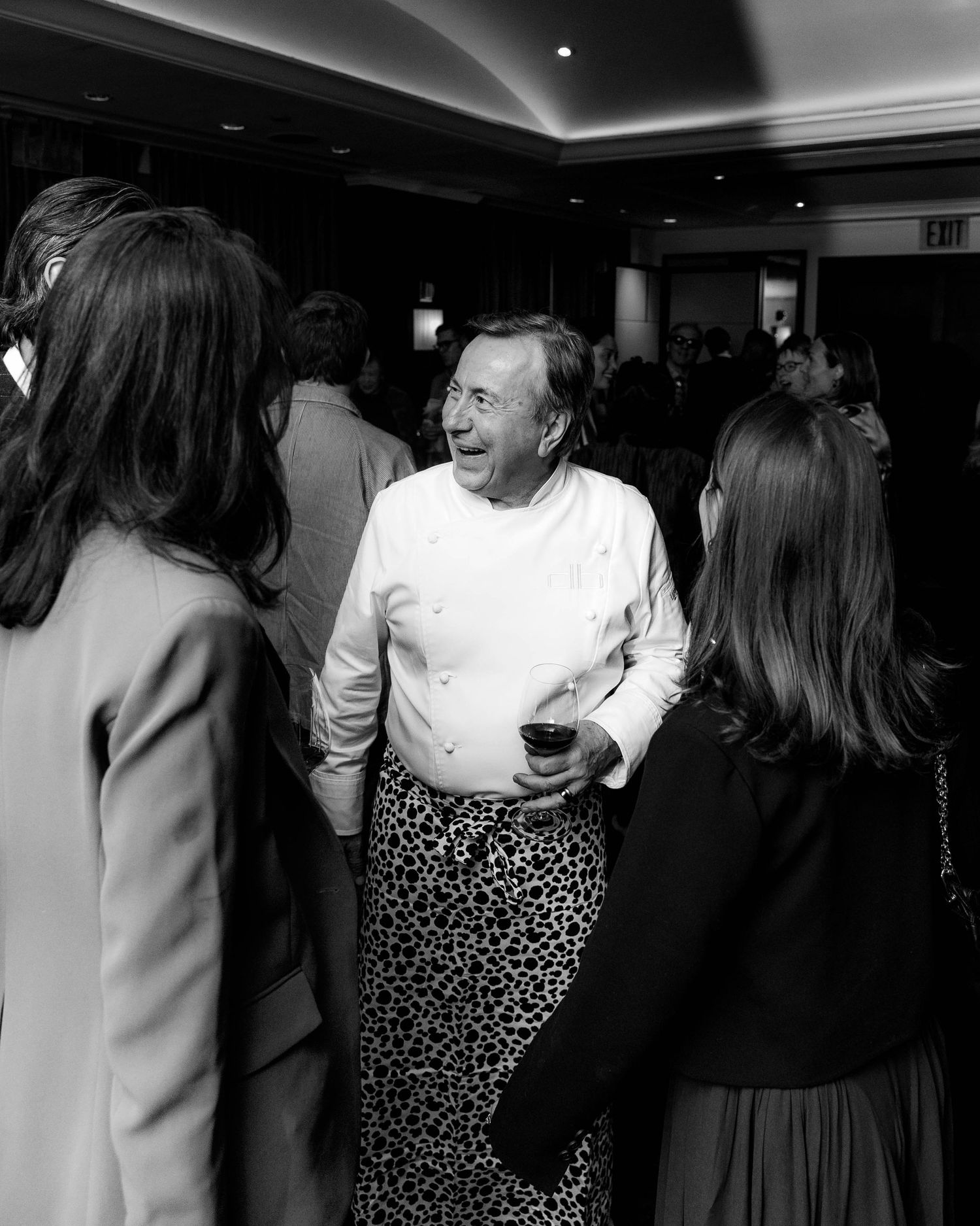 Black and white photo of a smiling man in a white chef's jacket and patterned apron holding a drink while chatting with guests at a crowded 