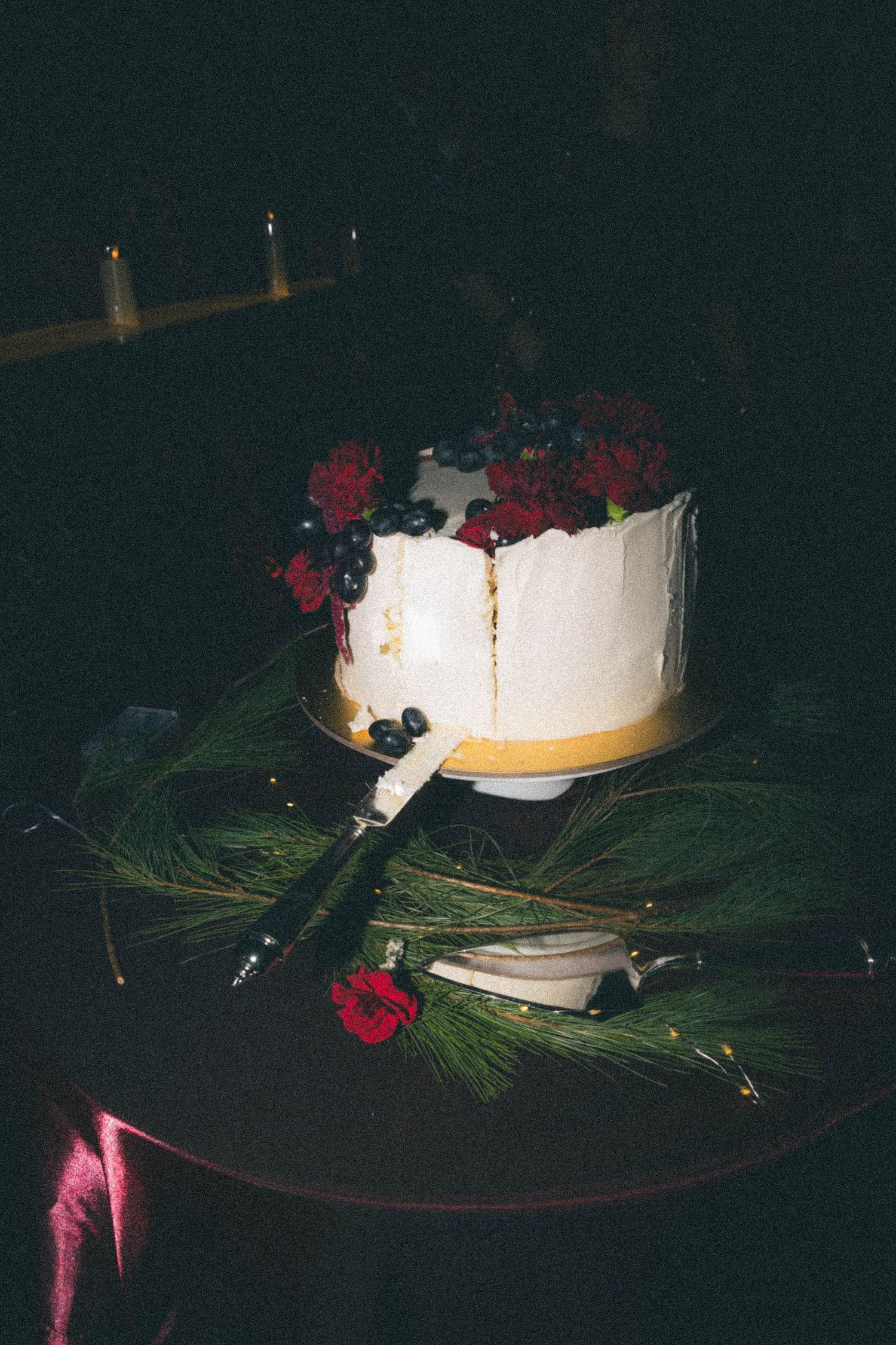 A white wedding cake topped with red flowers and blueberries on a decorated table at night.