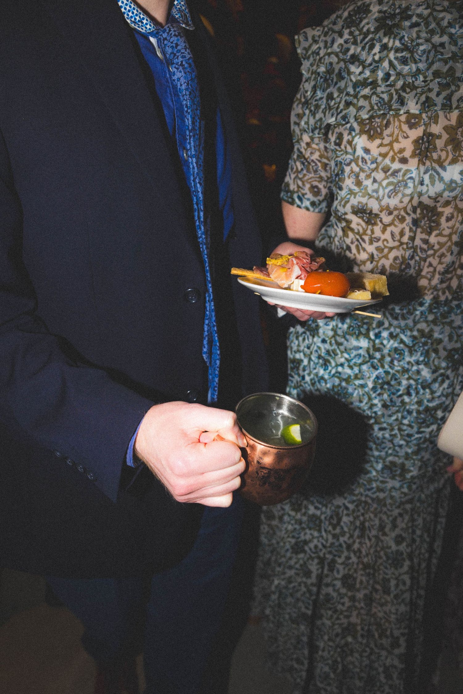 Wedding guest in navy suit holding a copper Moscow mule mug beside a woman with a food plate.