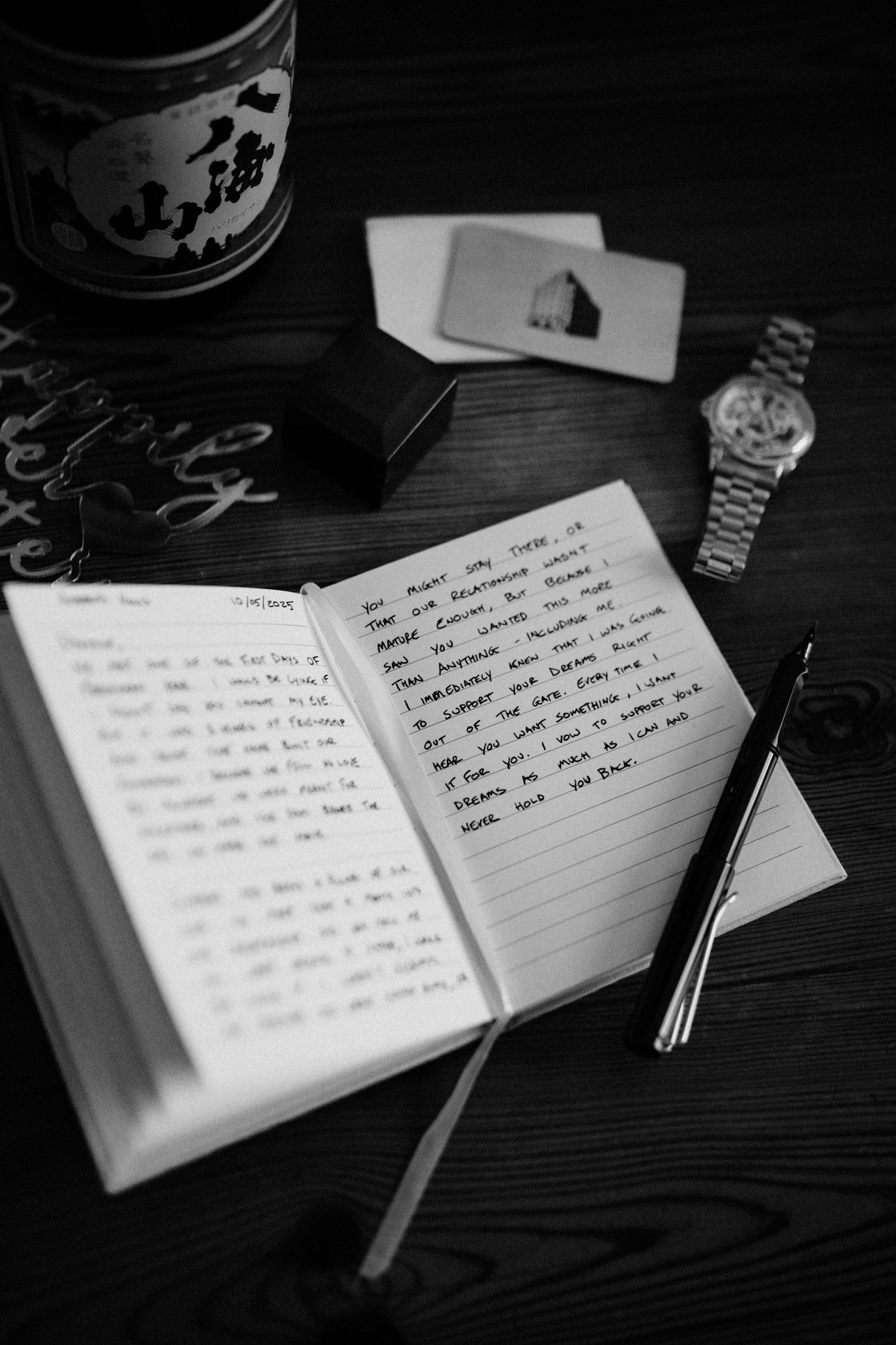 Black and white photo of an open handwritten journal beside a watch and sake bottle on a dark table.
