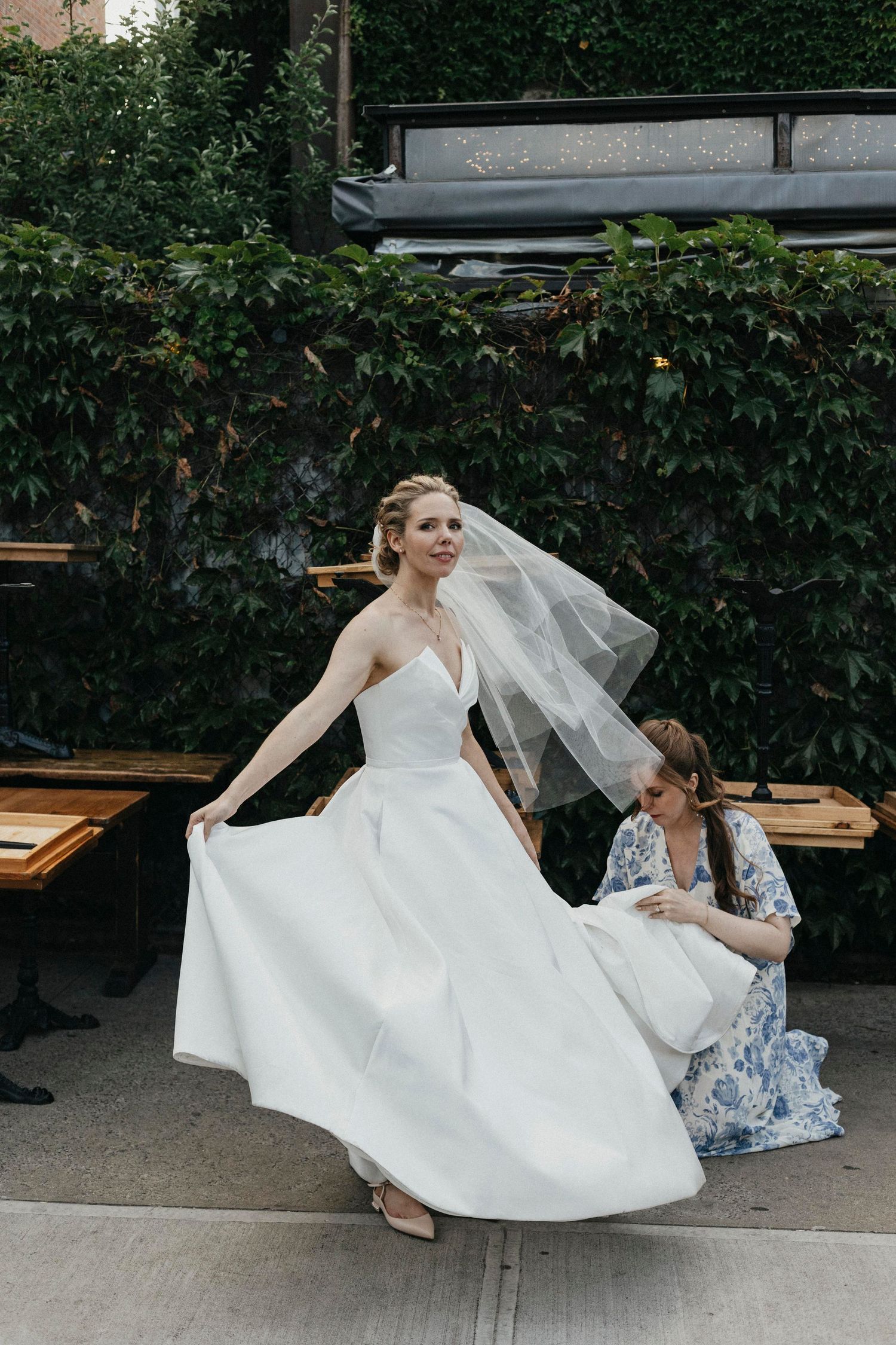 Bride in strapless white gown twirling her dress while bridesmaid in floral dress adjusts veil outdoors.