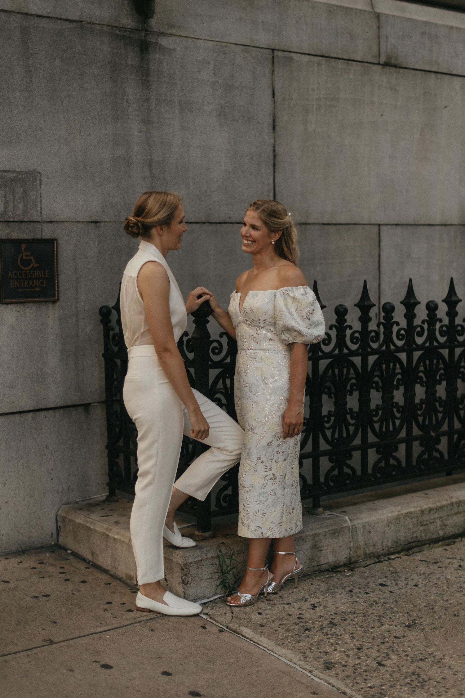 Two brides in white outfits laugh together outside a building with iron fence.
