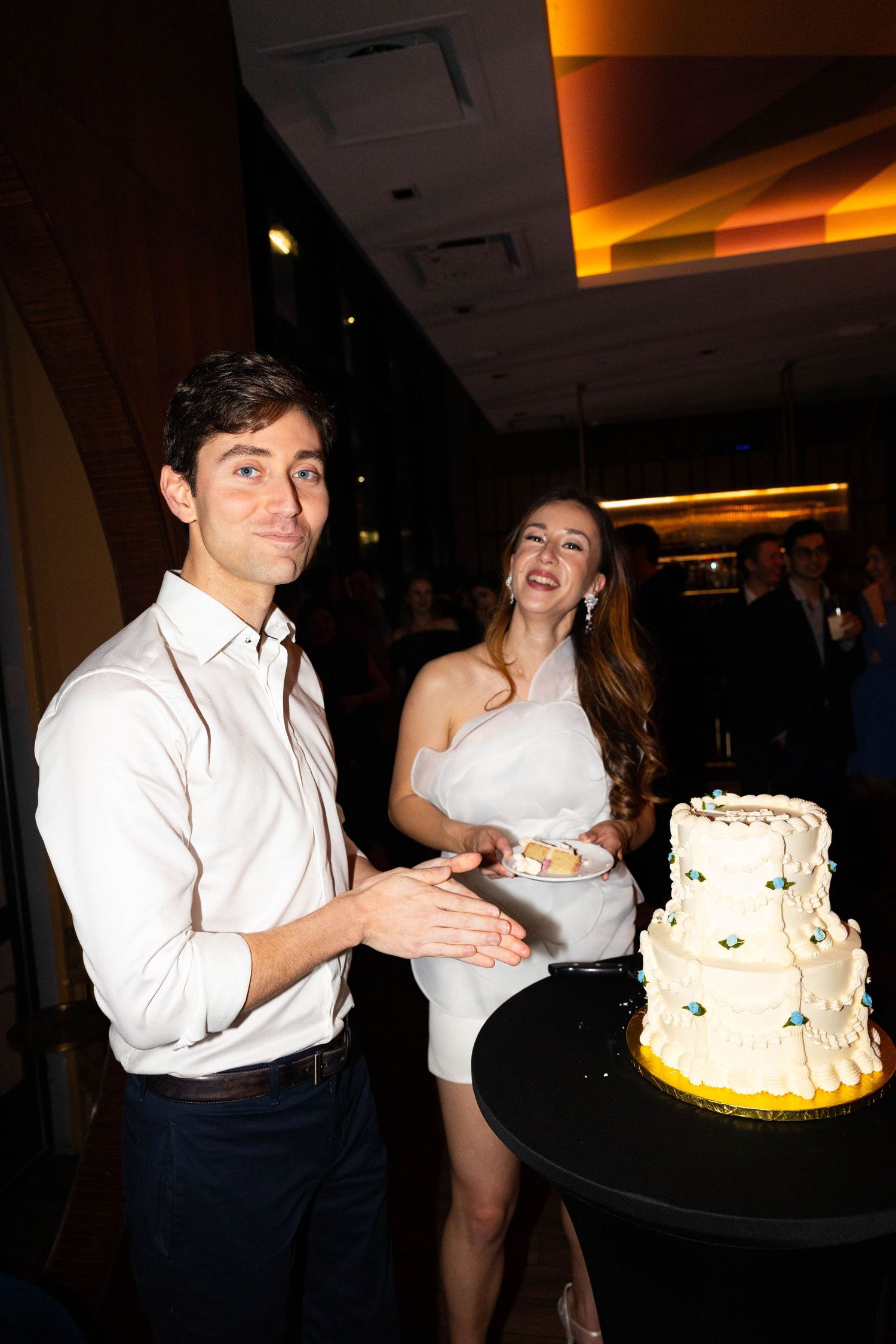 Smiling couple posing beside three-tier white wedding cake at dimly lit celebration venue.