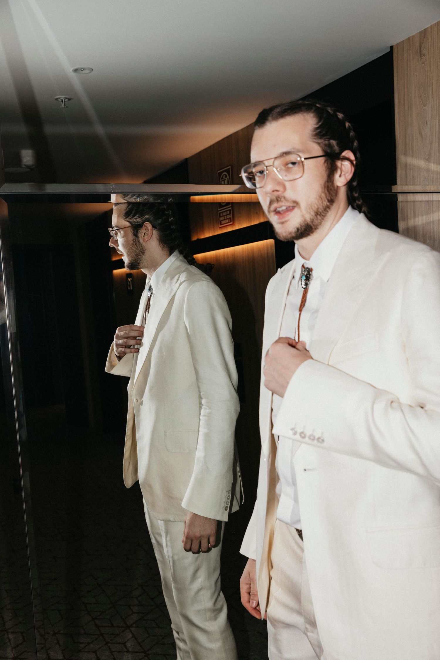 Groom in white suit with bolo tie checks his reflection in a mirror before his wedding.