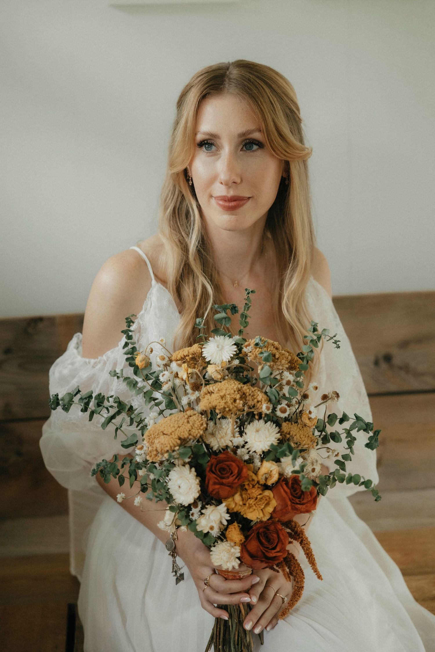 Blonde bride holds a lush bouquet of rust, white, and gold wildflowers in a bright studio.