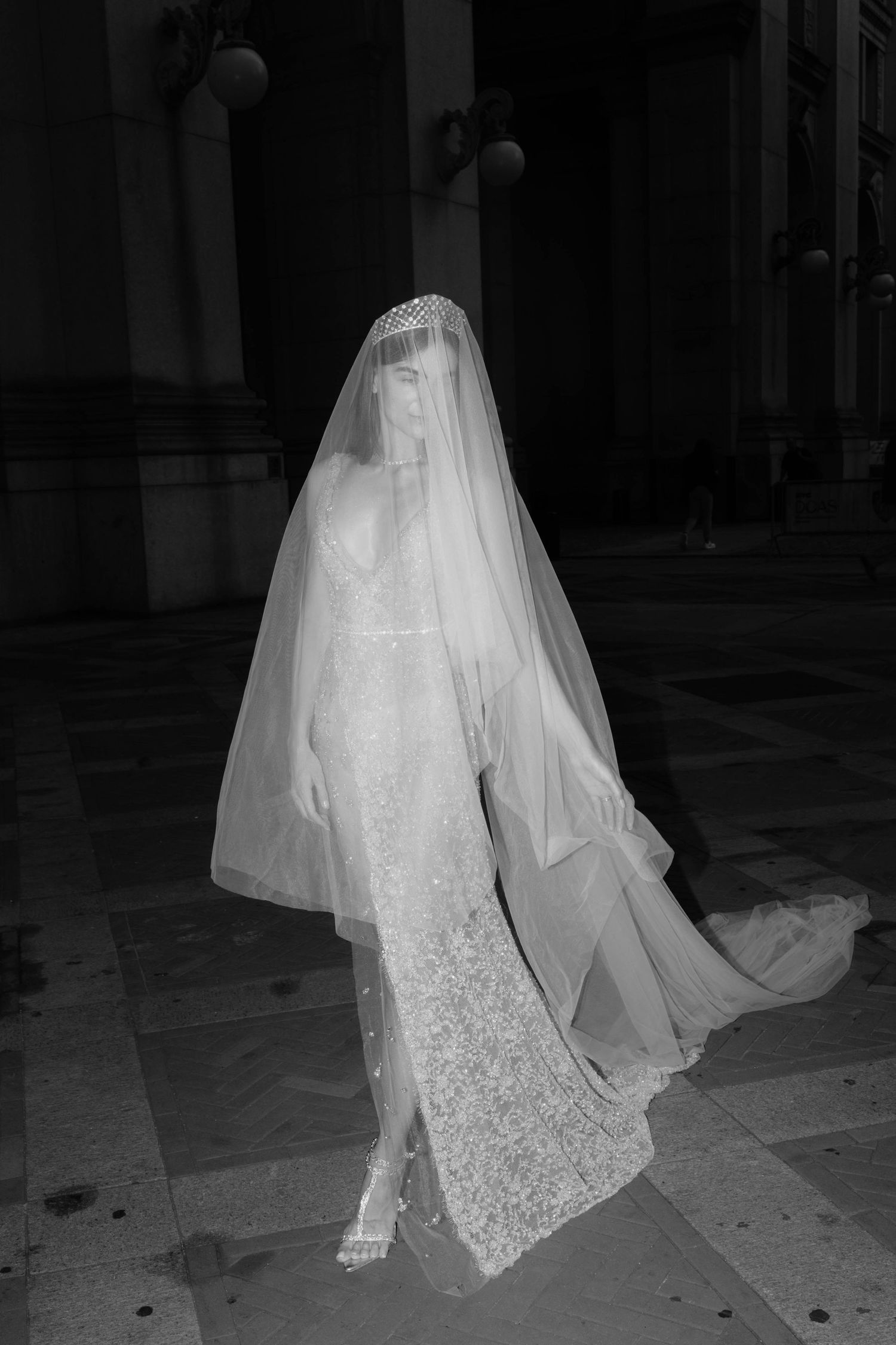 Black and white photo of bride in beaded gown with dramatic veil draped over face in grand interior.