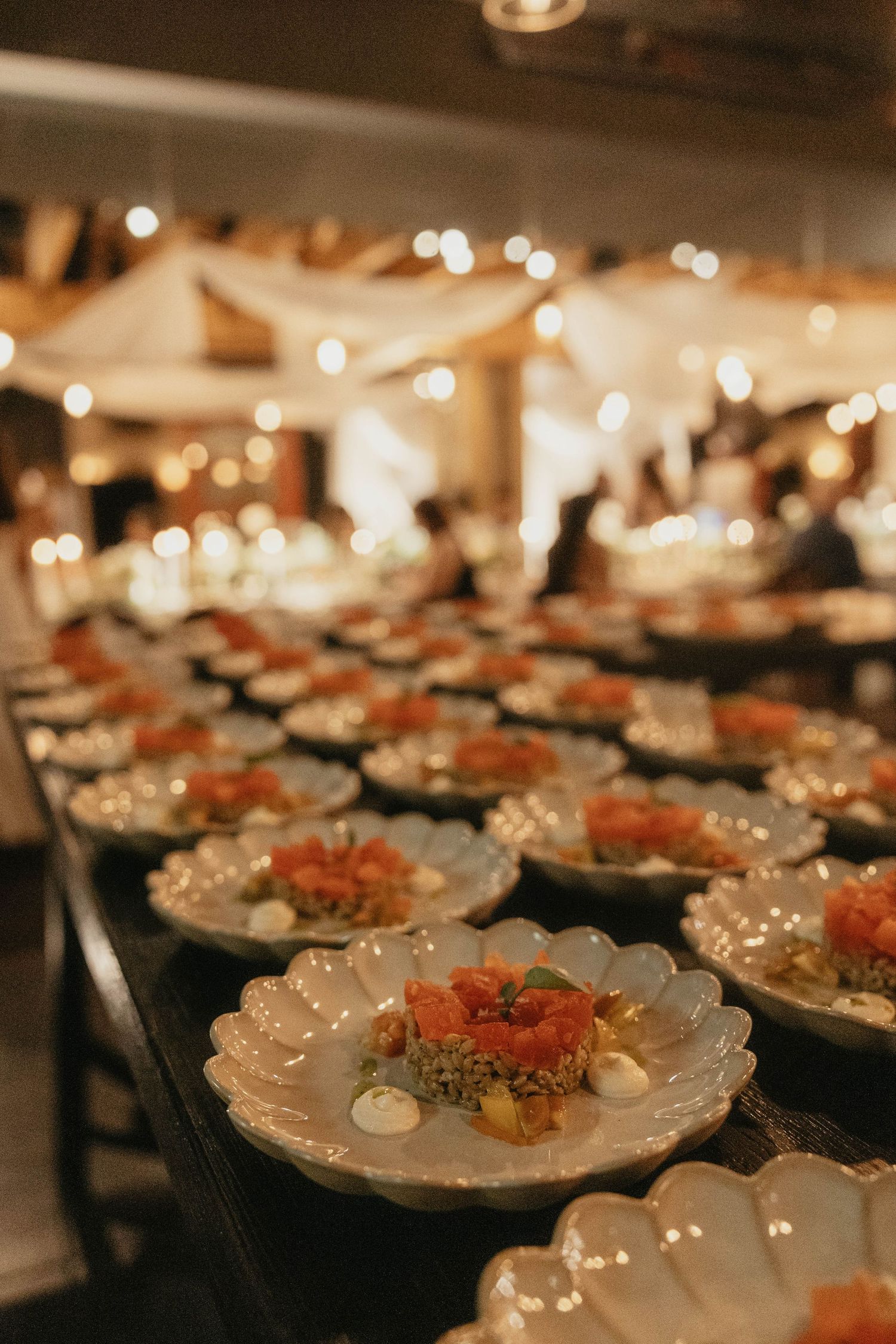 Row of scalloped plates topped with fresh tartare and creamy accents on a long banquet table at an elegant event with warm string lights.