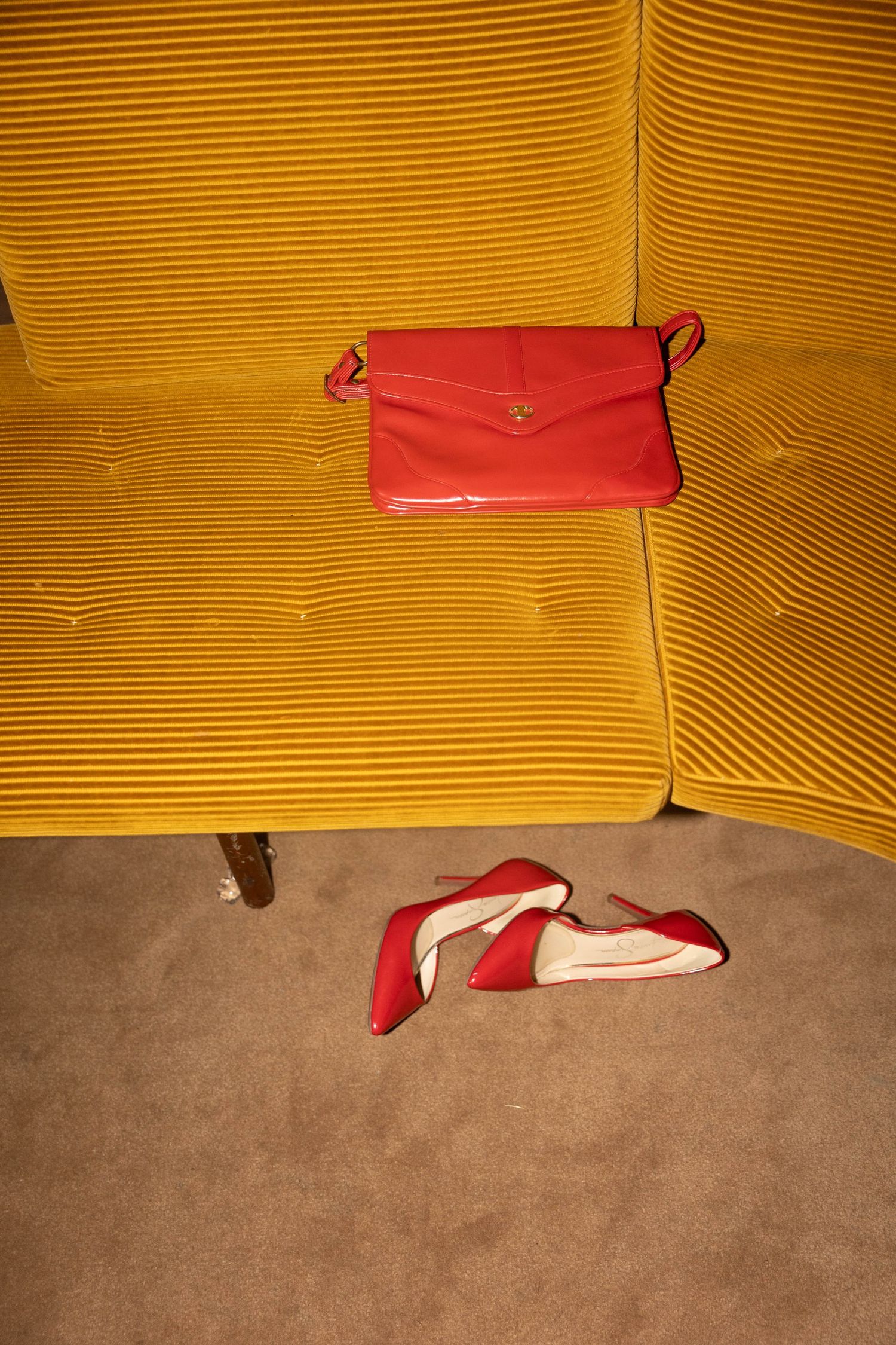 Red leather clutch bag on a mustard yellow corduroy sofa with a pair of red high heel pumps lying on the beige carpet below.