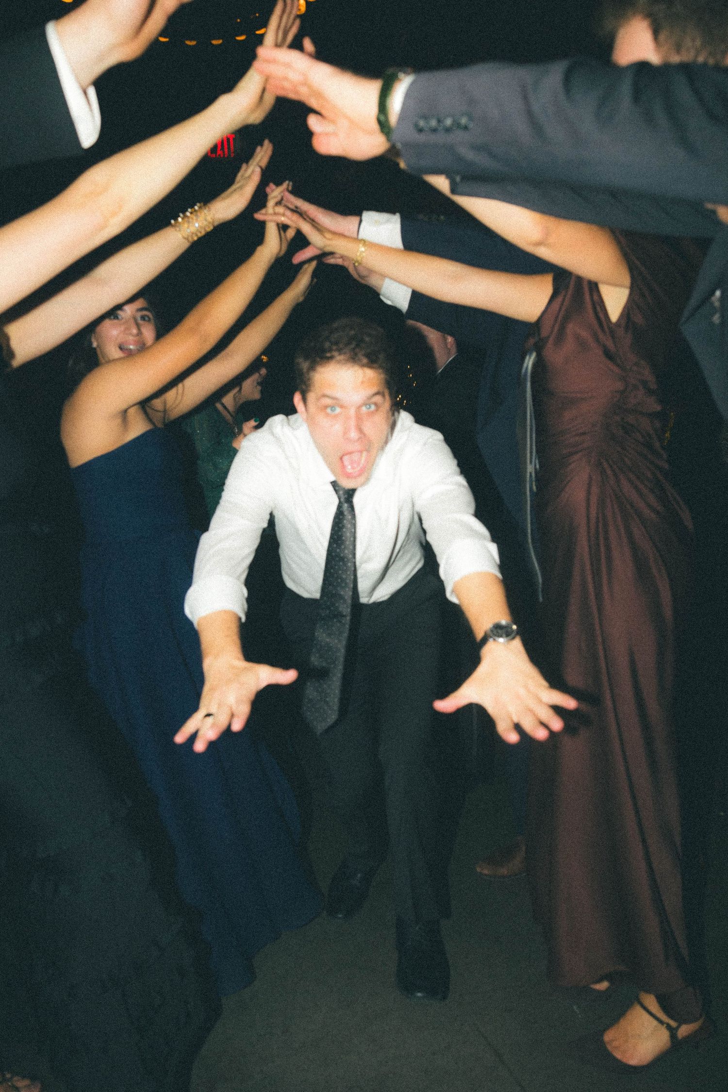 Man in white shirt and tie ducks under arch of raised arms at lively wedding reception dance floor.