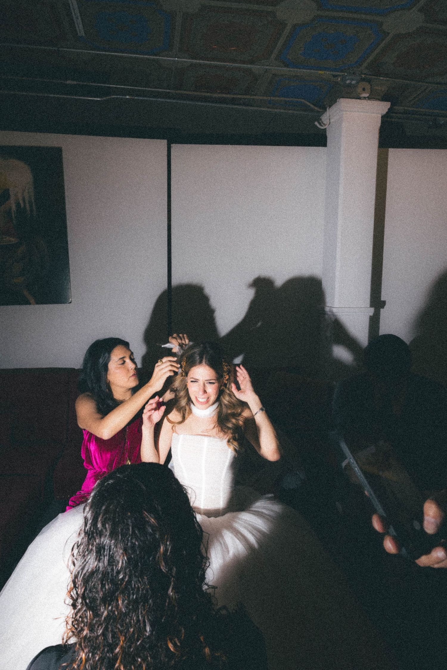 Bride in white dress gets hair adjusted by bridesmaid in pink dress in dimly lit venue room.