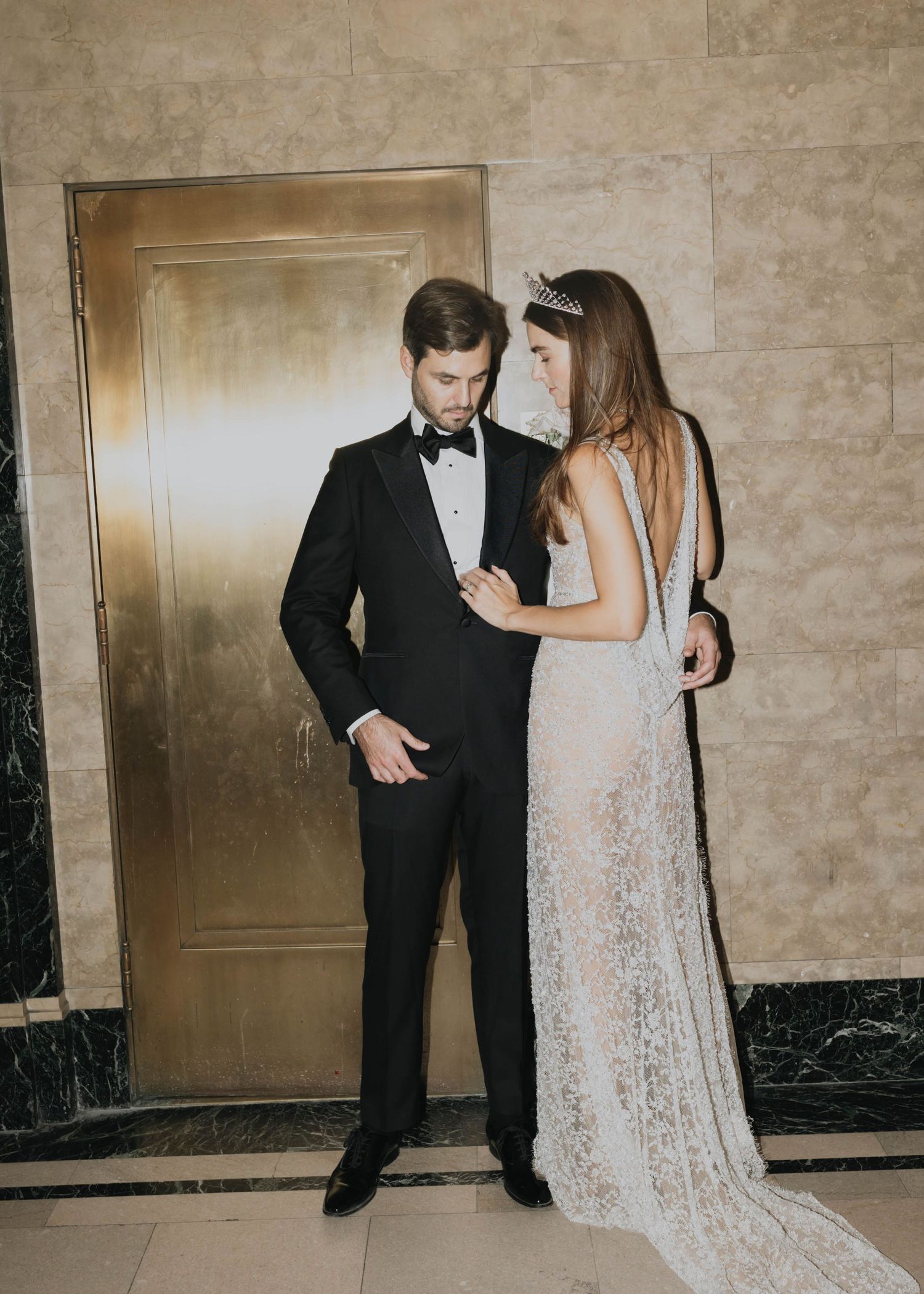 Bride in backless lace gown with tiara stands close to groom in black tuxedo near a gold elevator door.