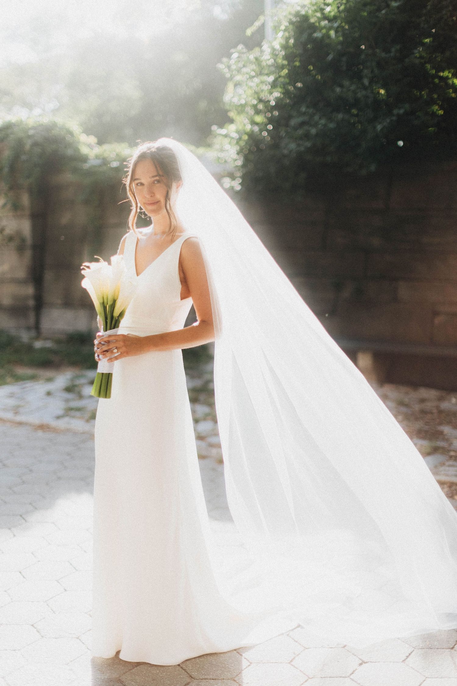 Elegant bride in white gown and flowing veil holding white calla lily bouquet in sunlit garden.