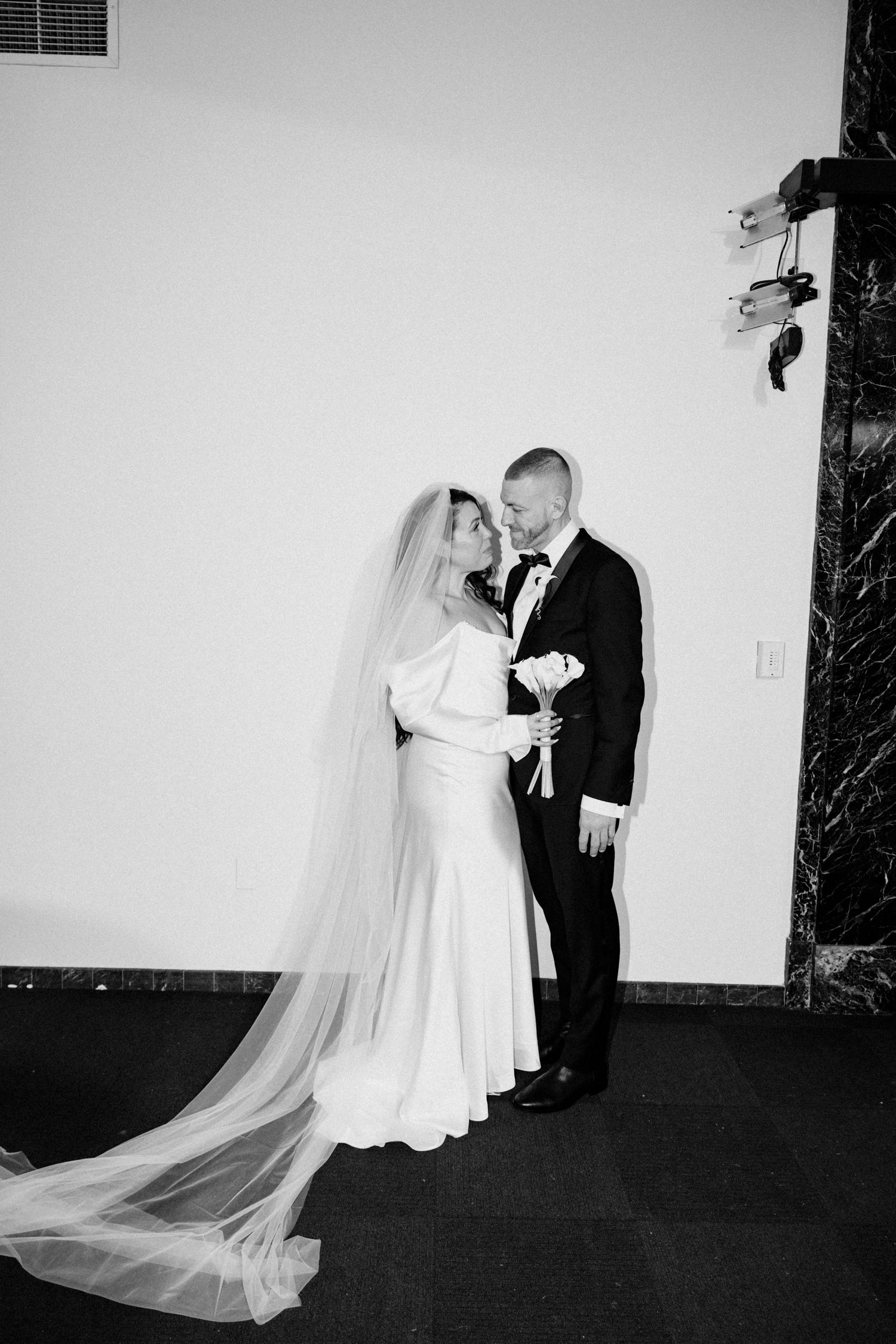 Bride in white gown with cathedral veil stands with groom in tuxedo, black and white.