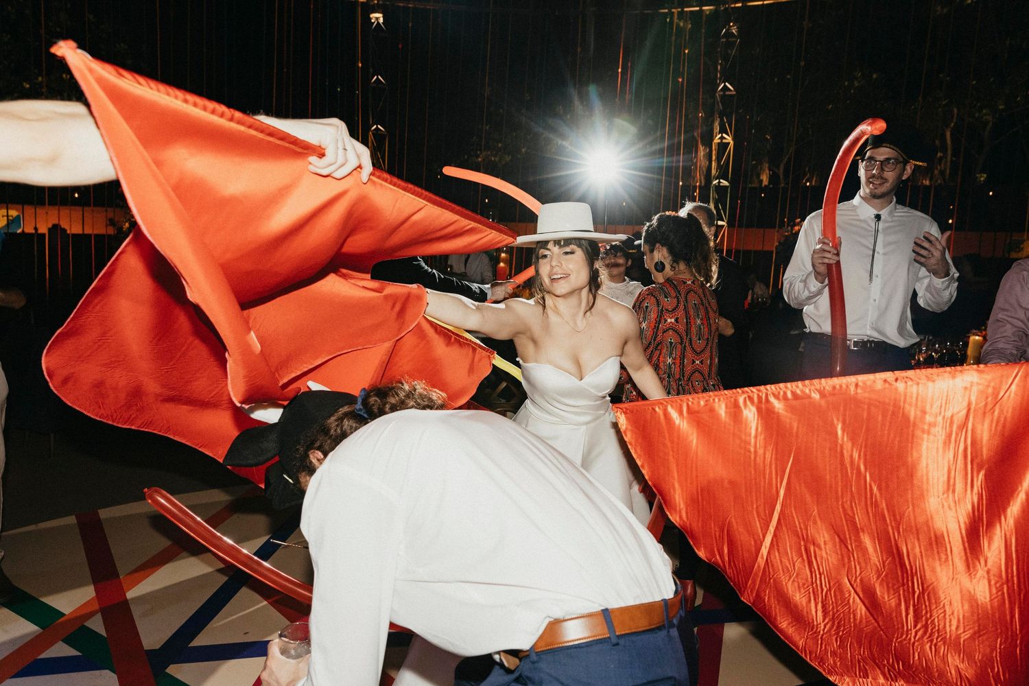 Bride in white gown and hat rides a mechanical bull with red cape at a festive wedding reception.