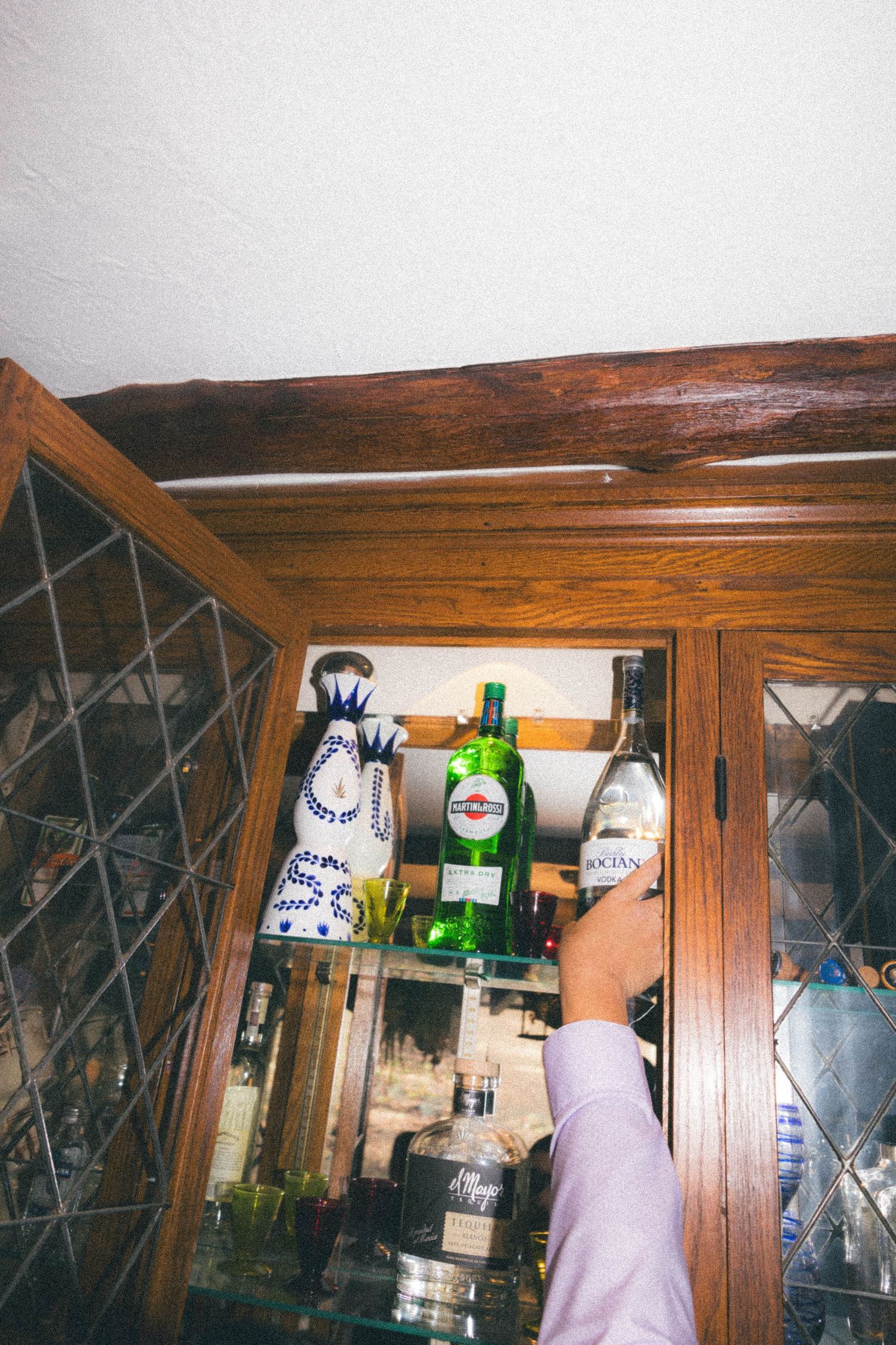 Hand reaching into a wooden bar cabinet stocked with bottles of Martini and other spirits.