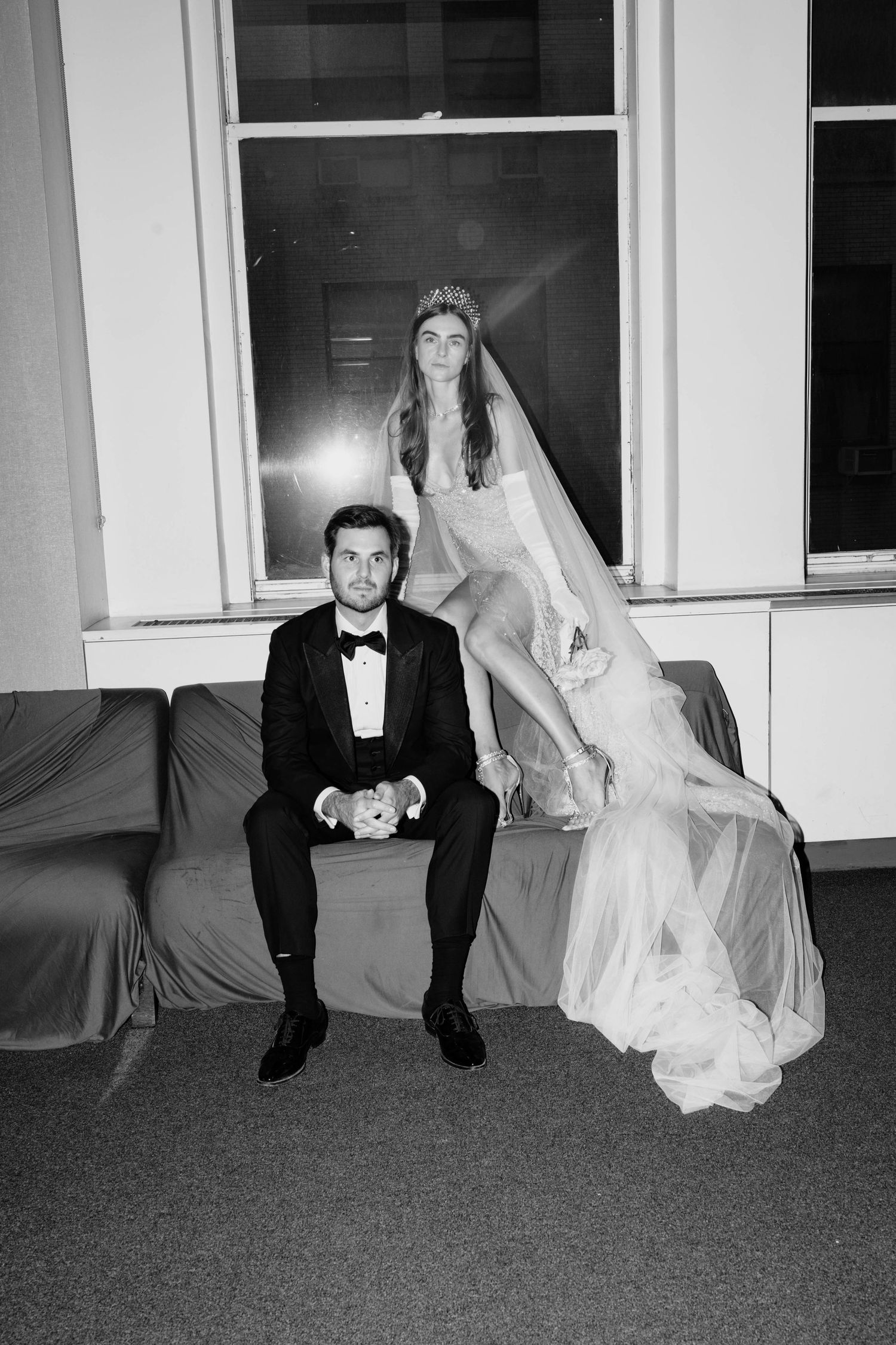 Black and white photo of bride in crown and groom in tuxedo posing on a sofa at their wedding reception.