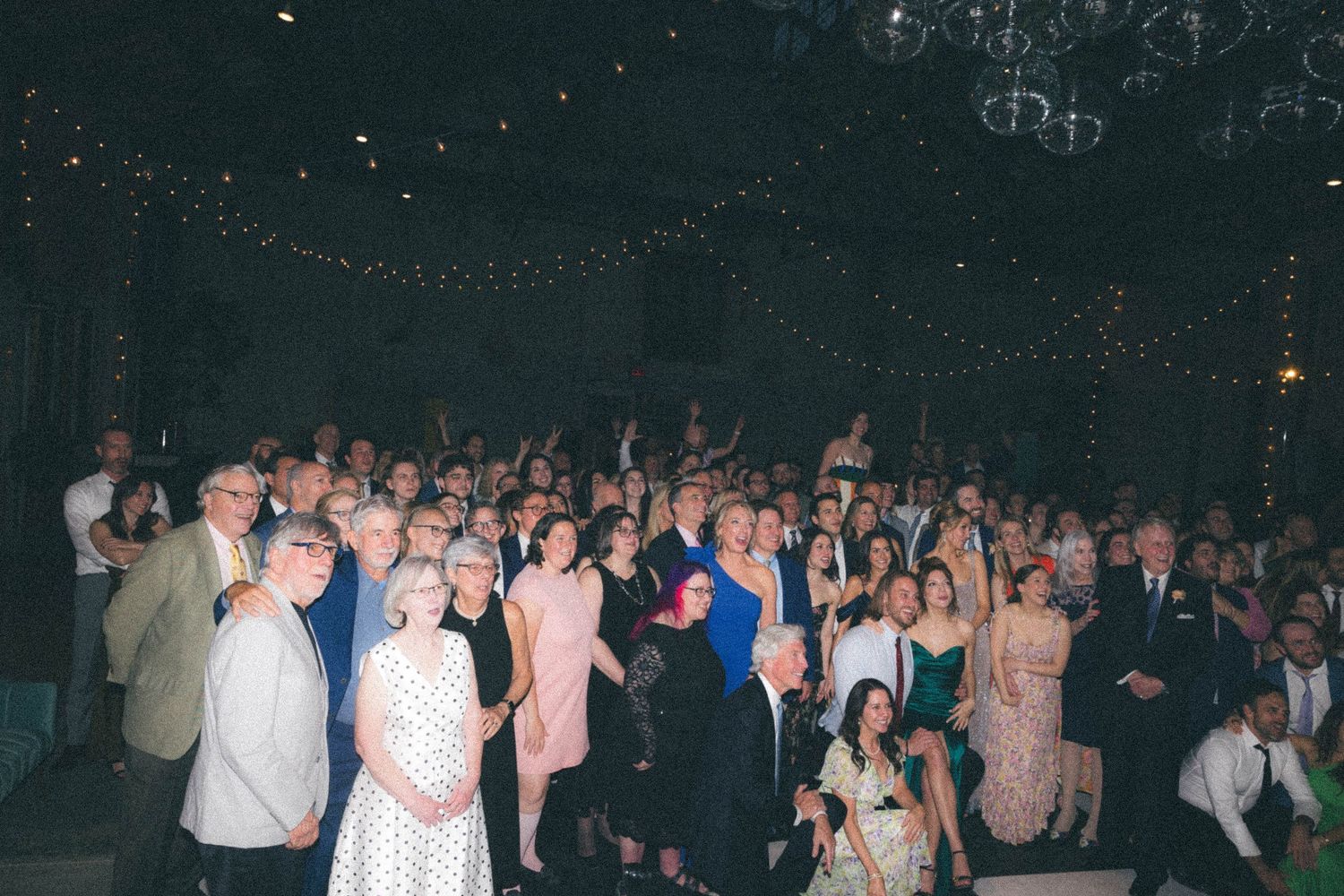 Large group of wedding guests pose together for a photo in a dimly lit reception venue.