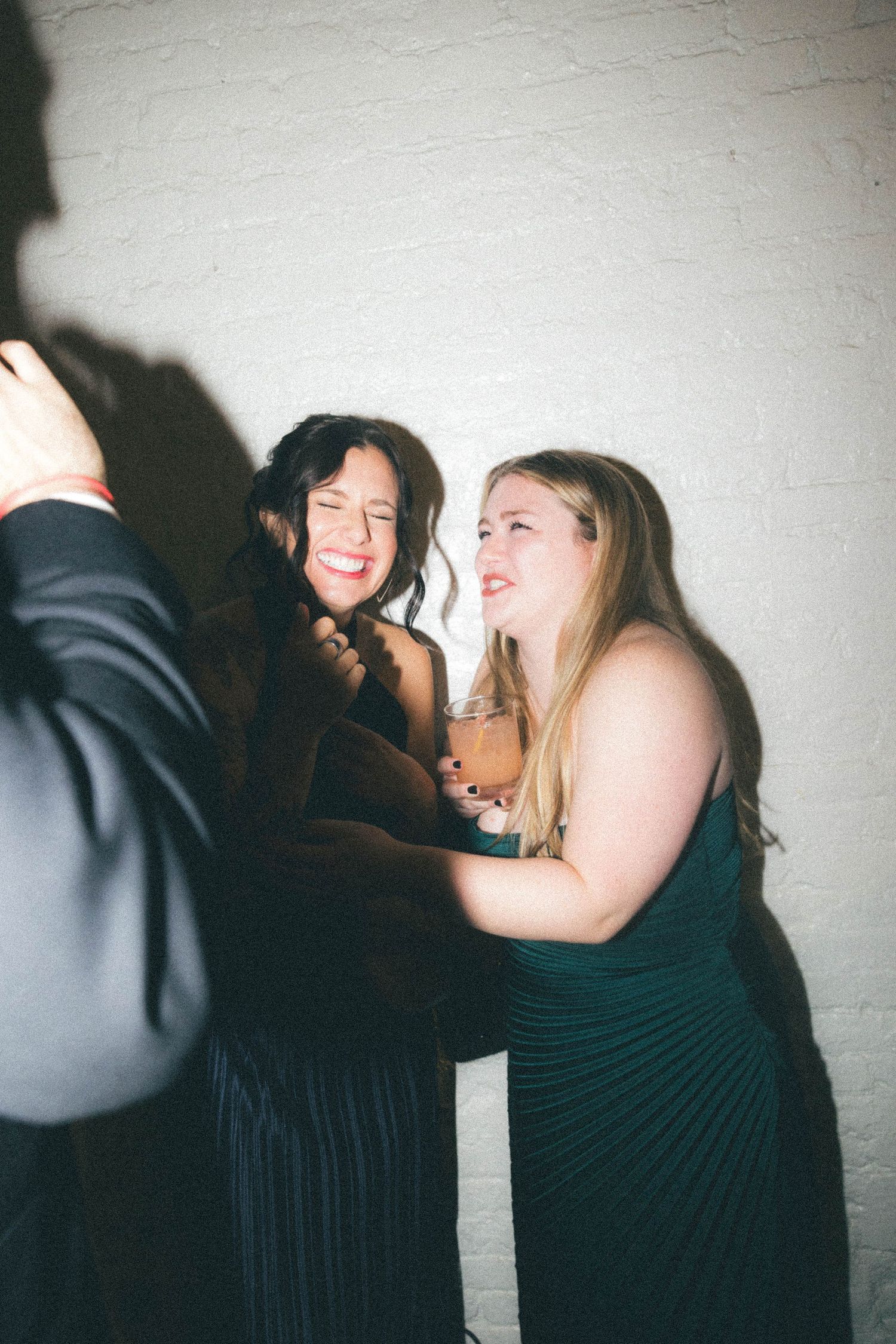 Two women laughing joyfully at formal evening event, one in black dress holding a cocktail glass.