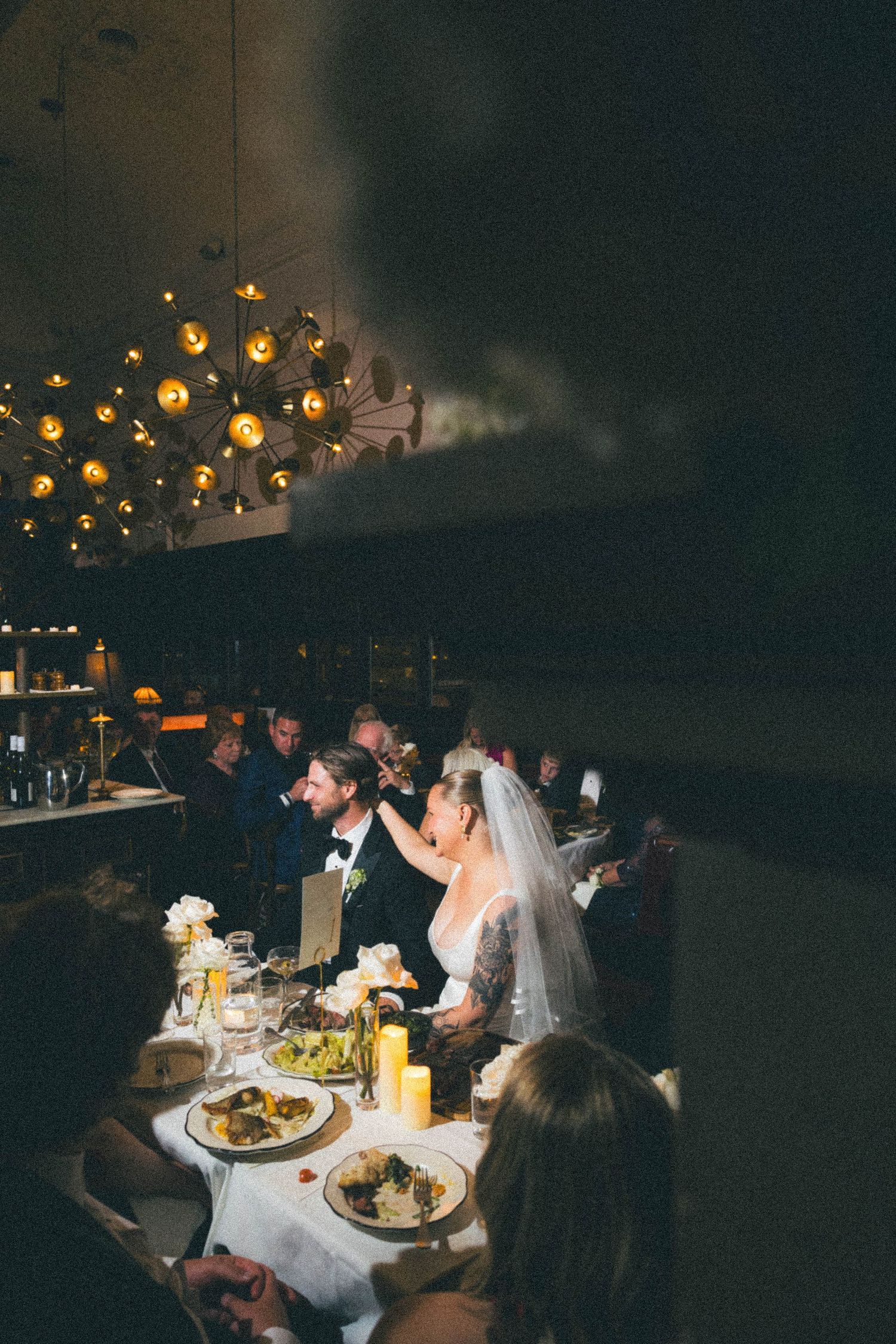 Bride and groom seated at candlelit wedding reception dinner table in warmly lit restaurant venue.