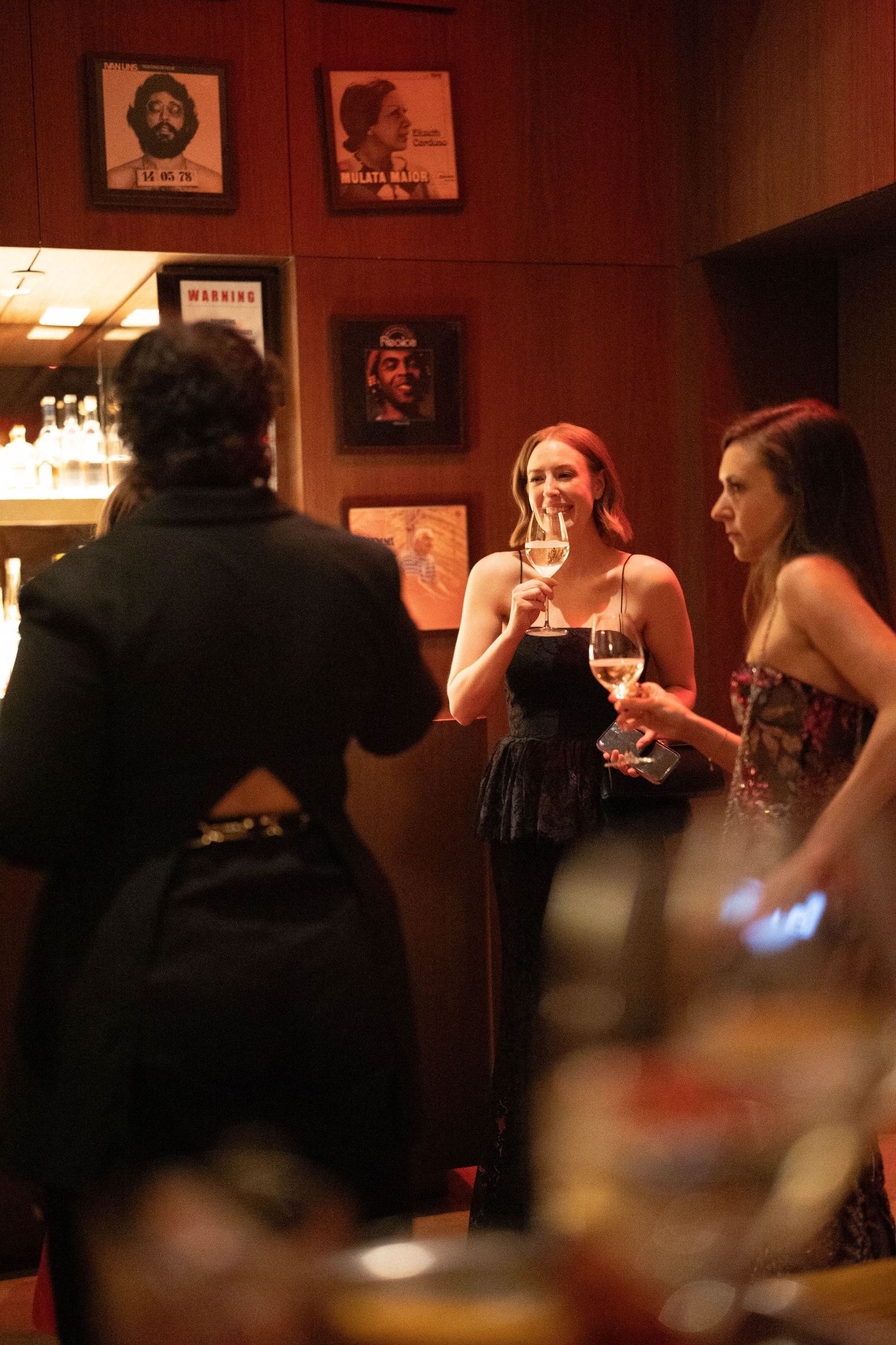 Three women in elegant evening attire chat and hold wine glasses in a dimly lit lounge with wood-paneled walls and framed portraits.