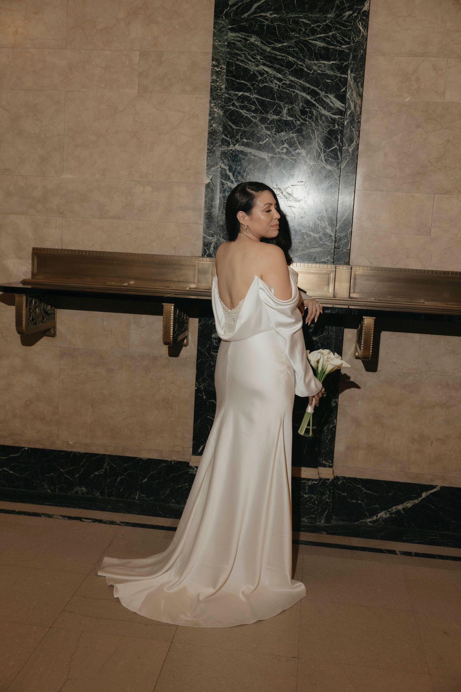 Elegant bride in off-shoulder satin wedding gown poses with bouquet near marble wall.