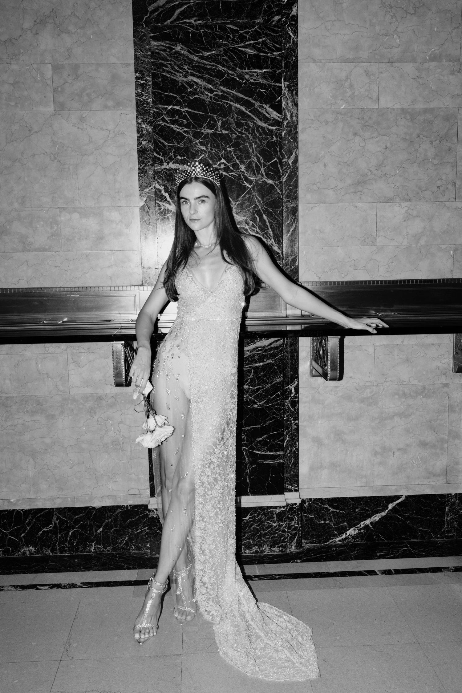 Black and white photo of bride in sheer lace gown with high slit and crystal crown posing elegantly.