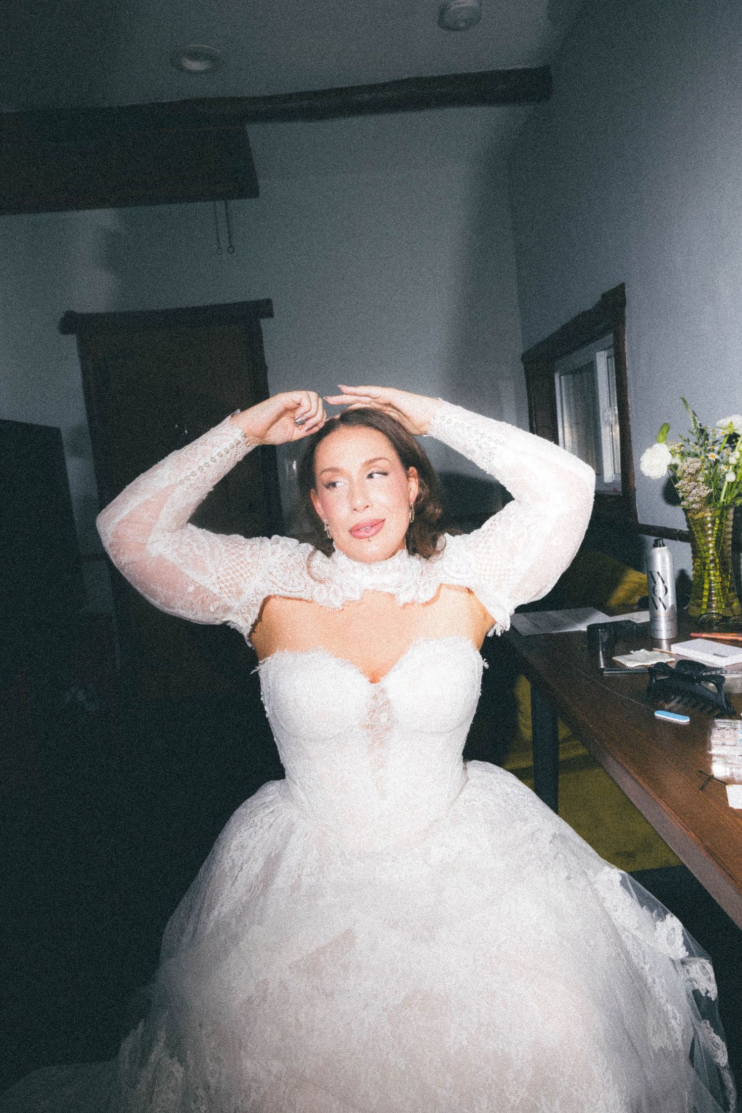 Bride in an elegant lace wedding gown adjusting her hair while sitting in a dimly lit room.