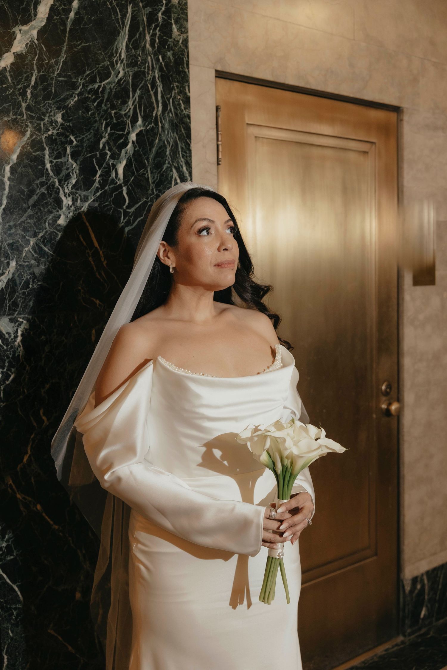Beautiful bride in off-shoulder white satin gown holding calla lily bouquet by marble wall.