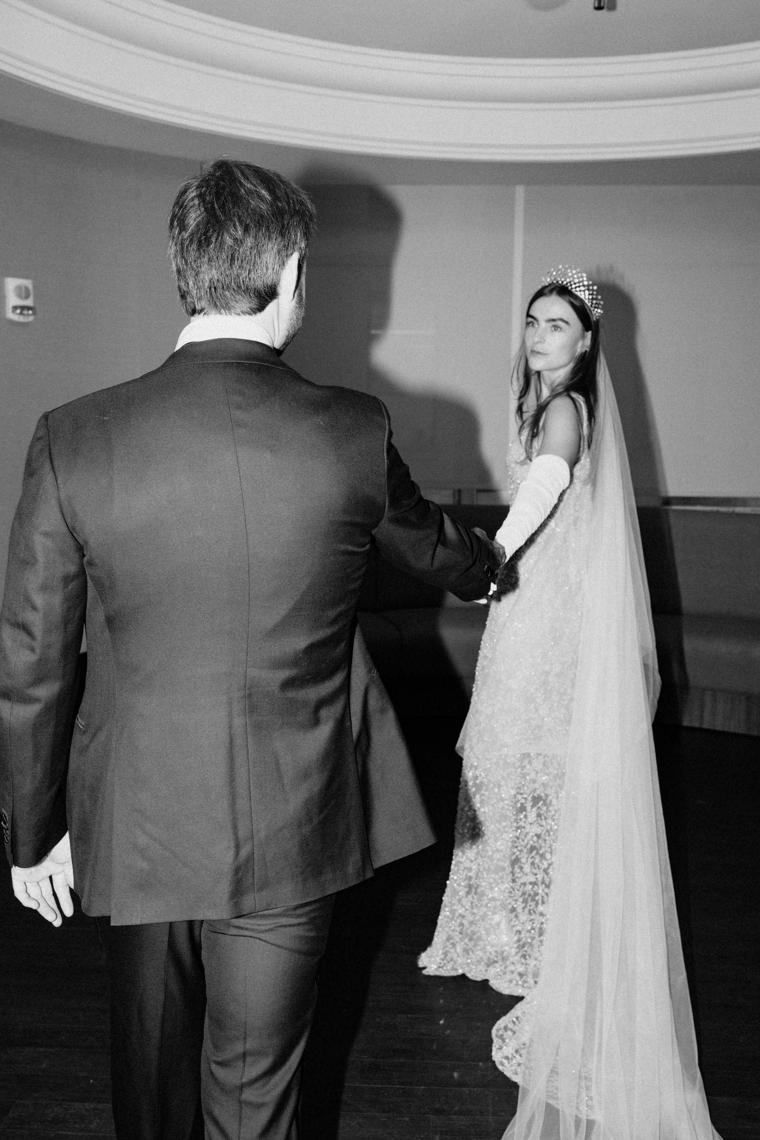 Black and white photo of bride in lace gown and crown holding grooms hand during first look moment.