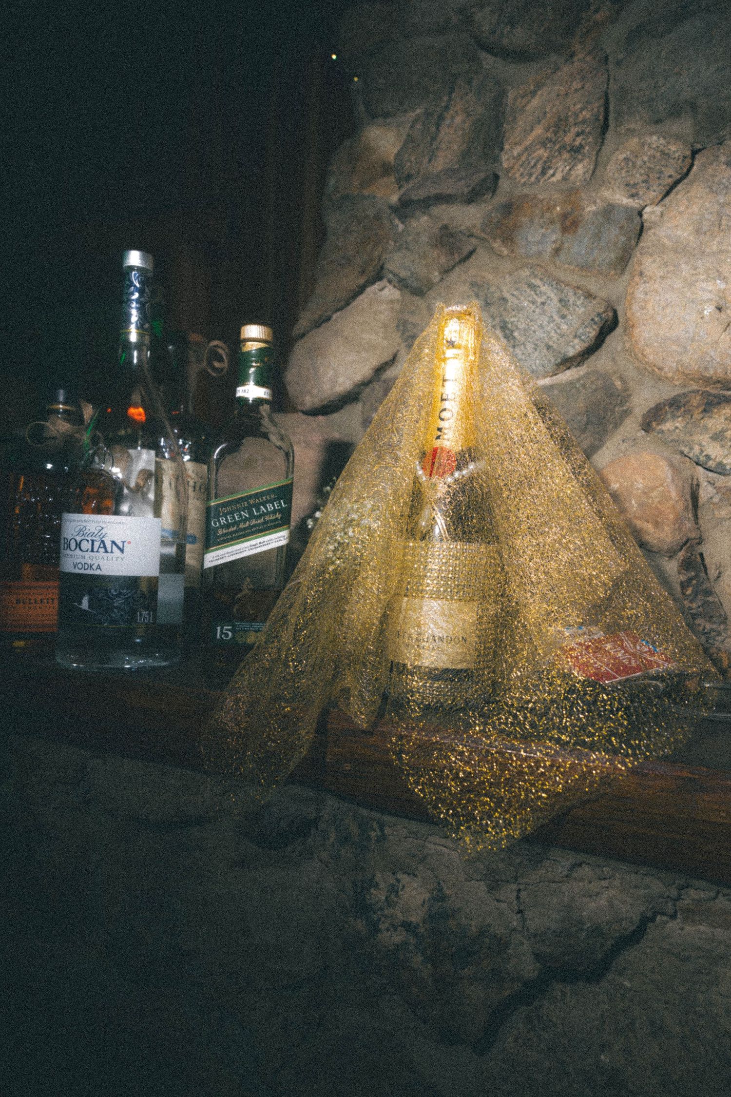 Champagne bottle wrapped in gold glittery pyramid display beside liquor bottles on stone wall.