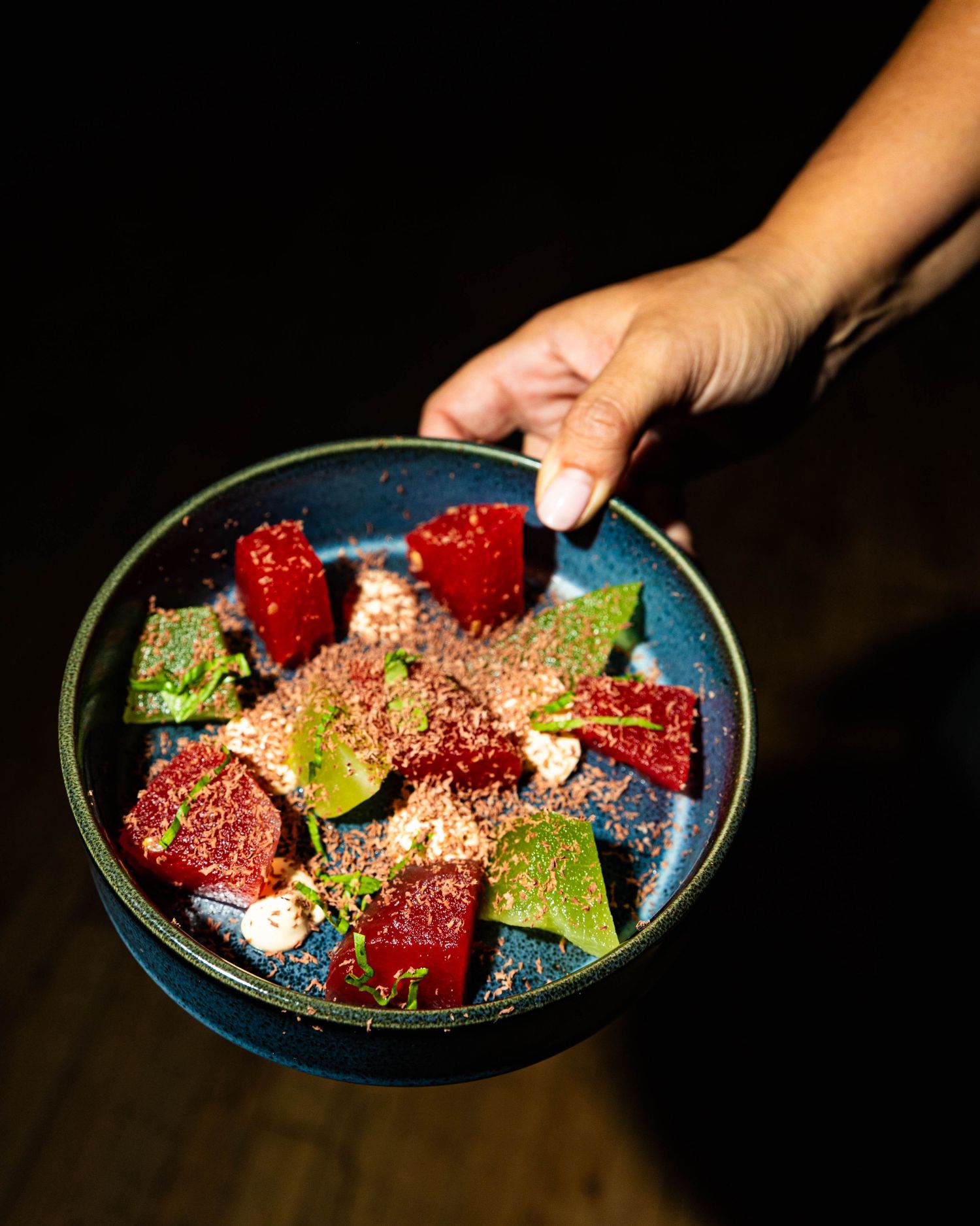 A hand holds a dark blue ceramic bowl of roasted beets with fresh green herbs, crumbled goat cheese and a sprinkle of toasted nut crumble.